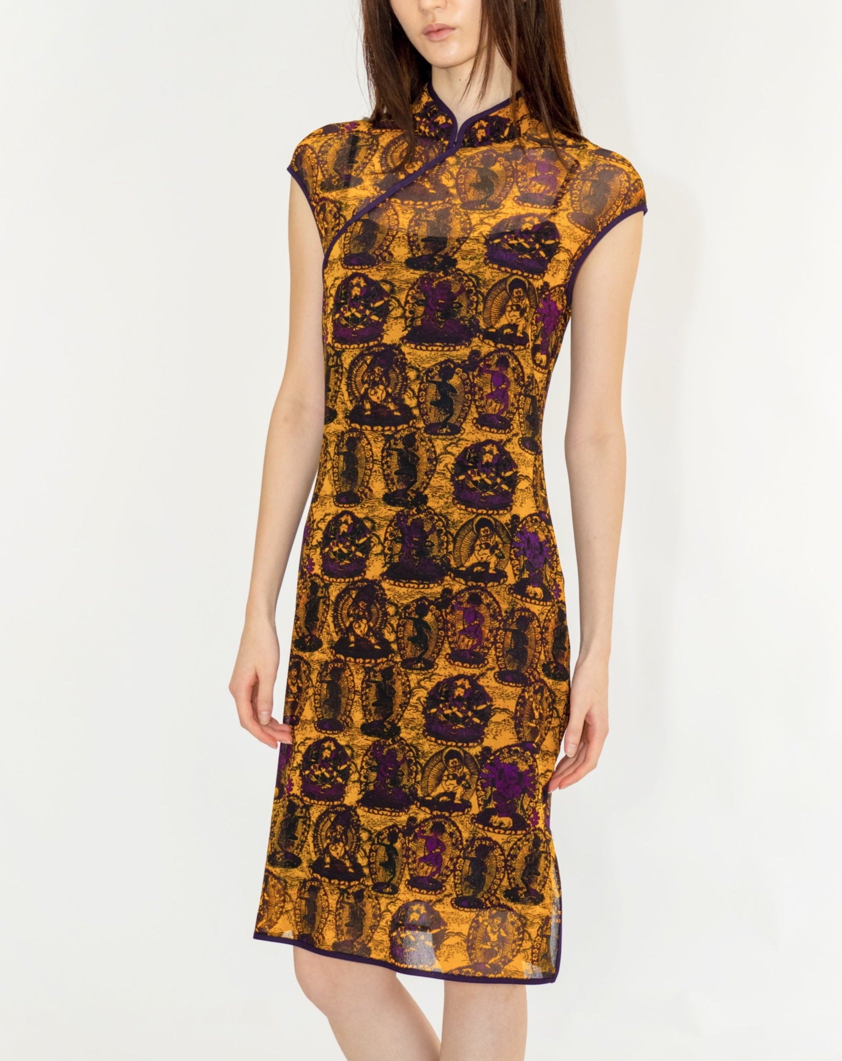 A woman modeling a sleeveless cheongsam-style dress with a dense, ornate pattern in gold, brown, and purple, paired with black loafers, against a plain white studio background.