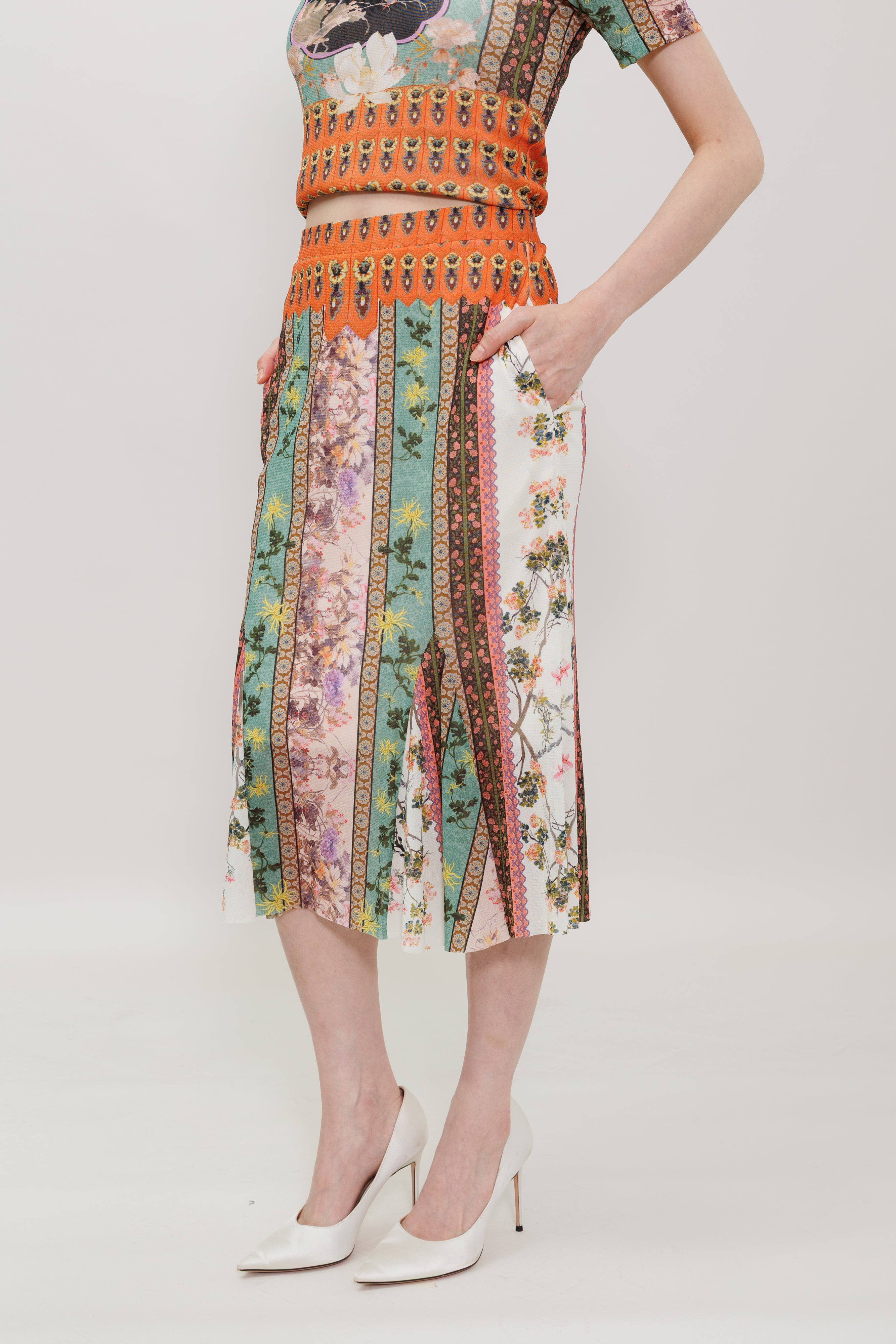 A model wearing a matching two-piece set featuring a cropped short-sleeve top and a pleated midi skirt, both with a vibrant multicolor vertical stripe pattern. The set includes orange, green, pink, and brown stripes with floral and geometric details.