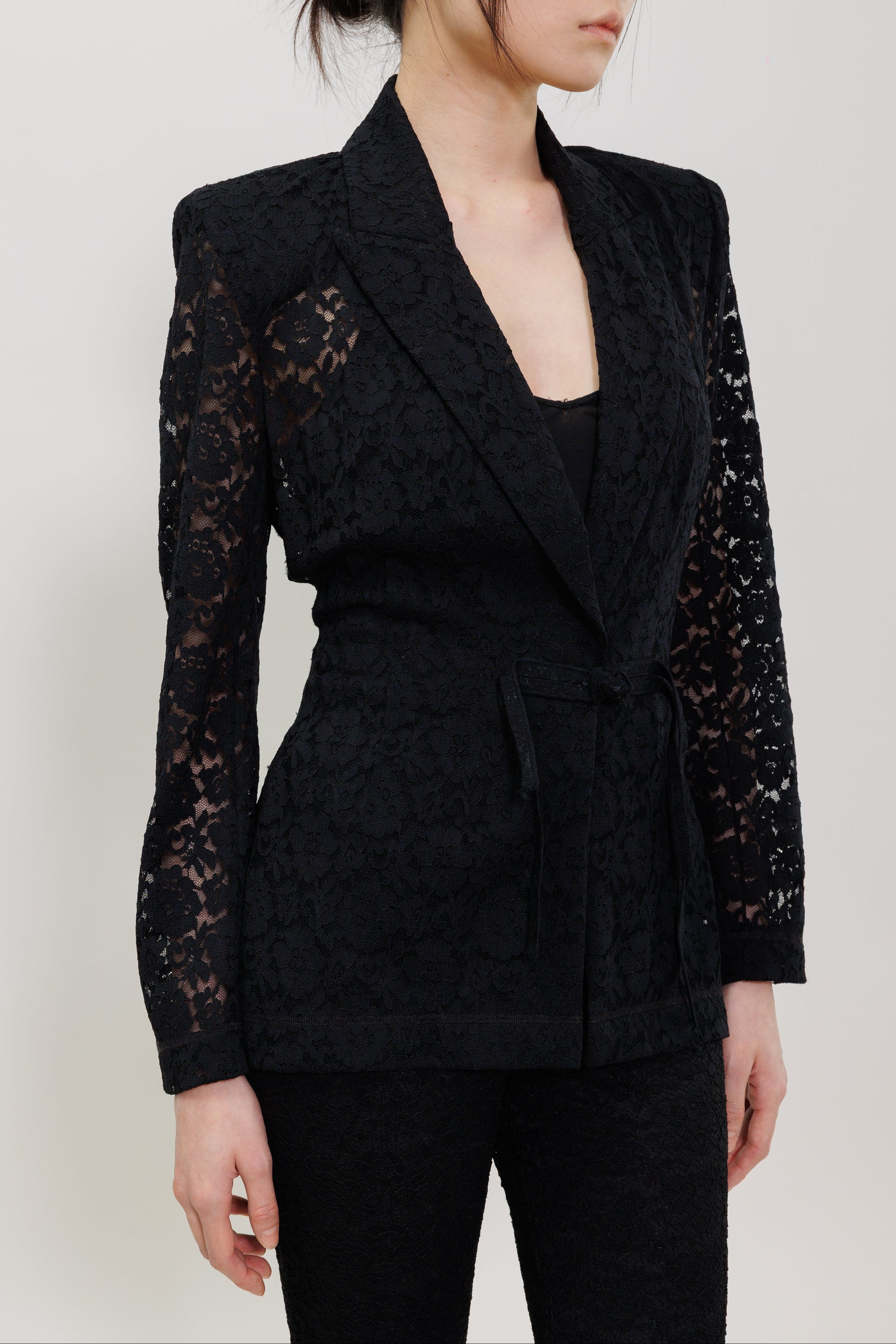 A woman wearing a black lace and textured fabric wrap-style blazer with long sheer lace sleeves, paired with black pants, photographed against a plain light gray studio background.