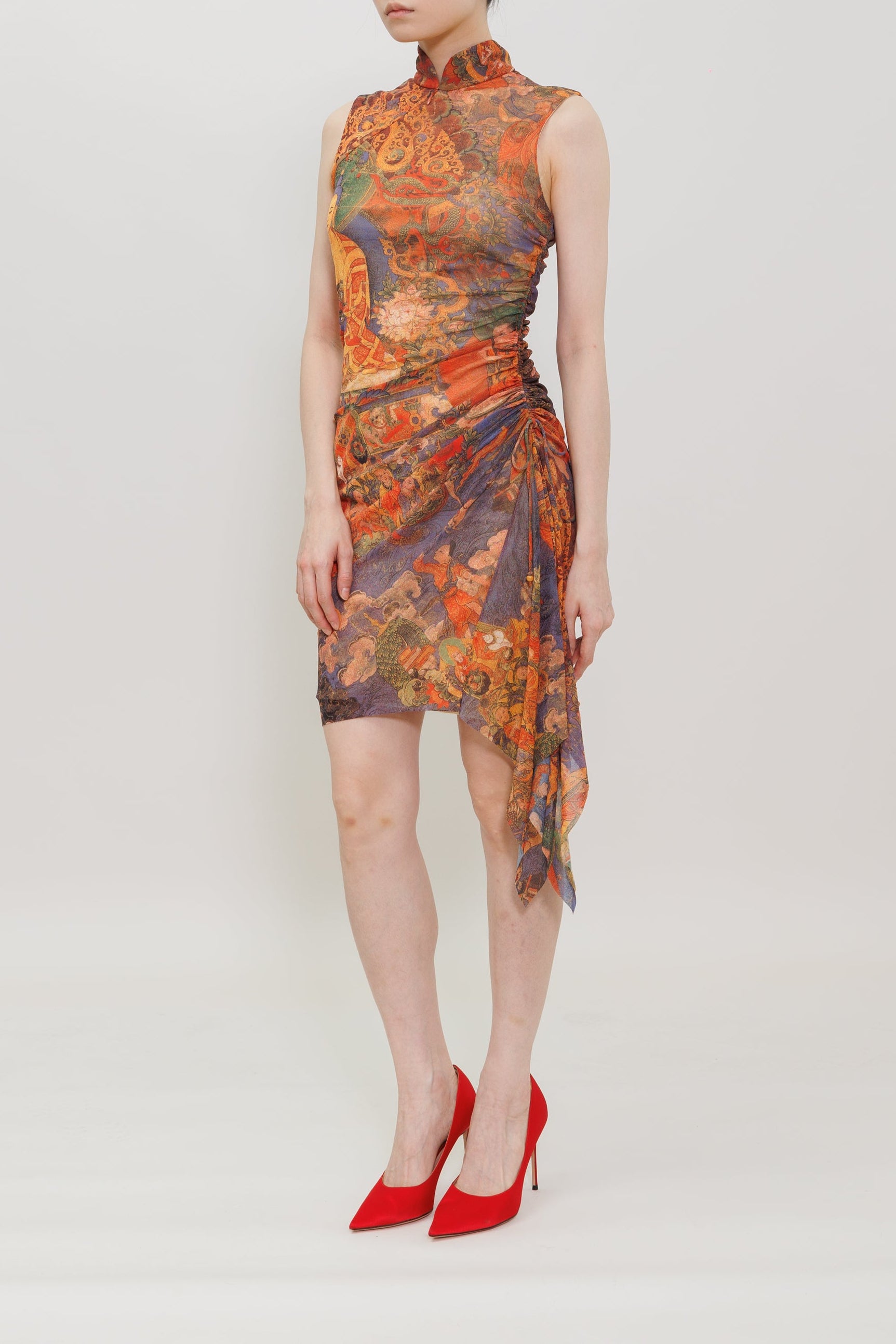 A model wearing a sleeveless high-neck mini dress with a vibrant orange, red, and purple abstract print, paired with red pointed-toe heels, against a plain light gray studio background.