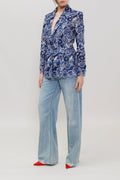 A model stands against a light gray studio background wearing a blue and white patterned button-up blazer, light-wash wide-leg jeans, and red pointed shoes.