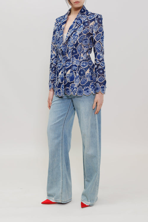 A model stands against a light gray studio background wearing a blue and white patterned button-up blazer, light-wash wide-leg jeans, and red pointed shoes.