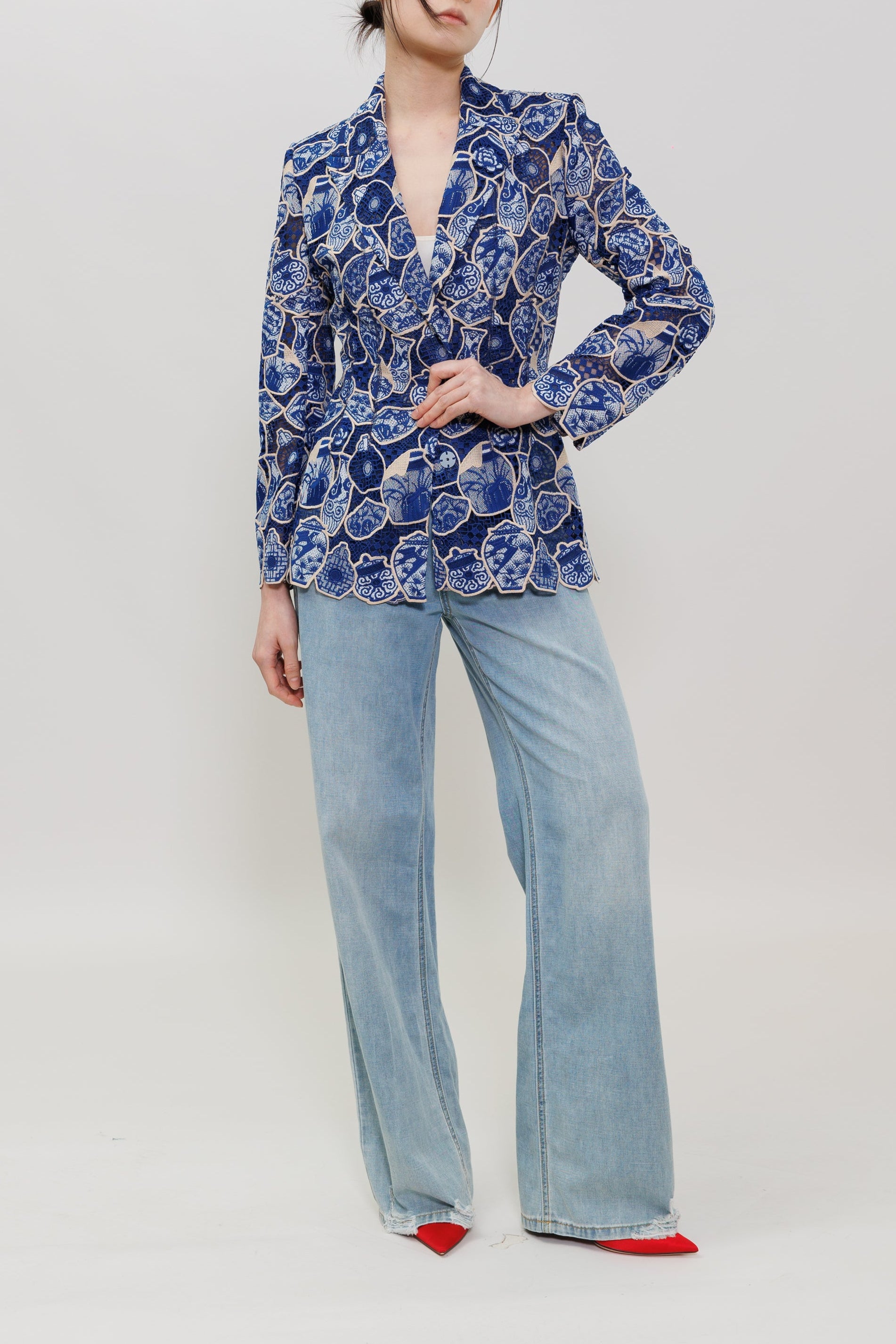 A model stands against a light gray studio background wearing a blue and white floral patterned blazer, light-wash wide-leg jeans, and red pointed shoes.