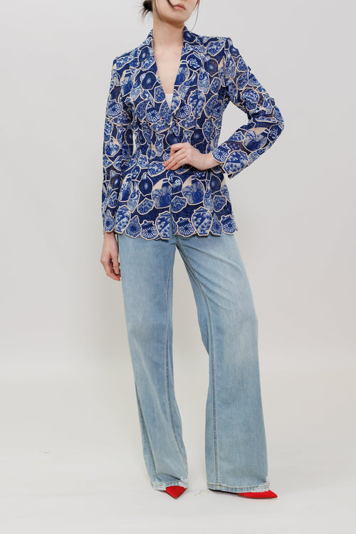 A model stands against a light gray studio background wearing a blue and white floral patterned blazer, light-wash wide-leg jeans, and red pointed shoes.