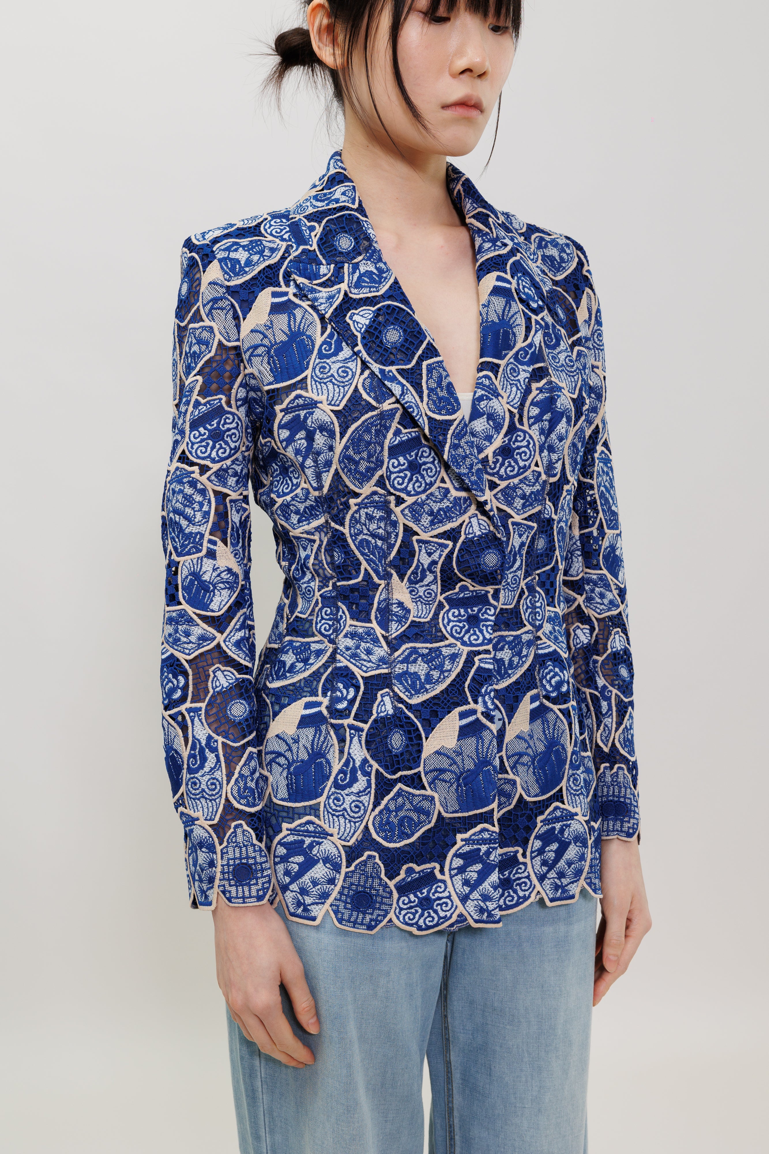 A woman wearing a blue and white floral print blazer with a deep V-neck and light blue jeans, photographed against a plain light gray studio background.