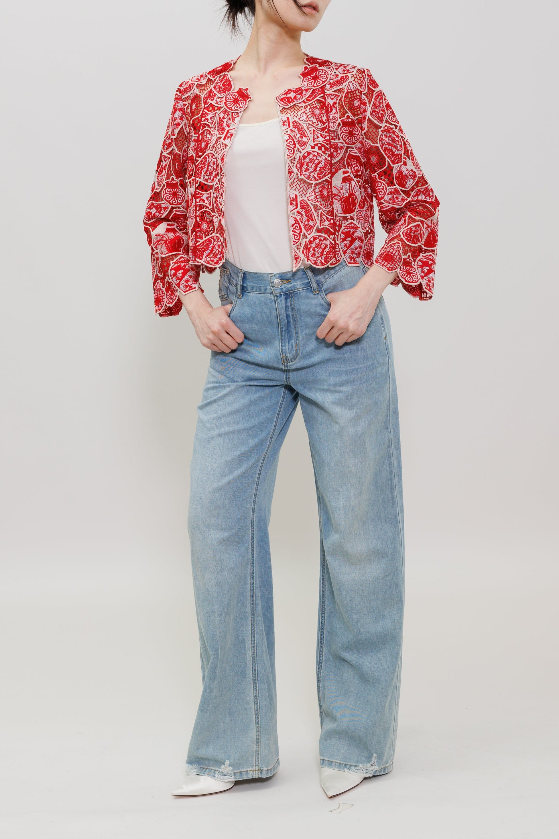A woman modeling a red and white floral patterned cropped jacket over a white top, paired with light blue wide-leg jeans and white pointed shoes, against a plain light gray studio background.