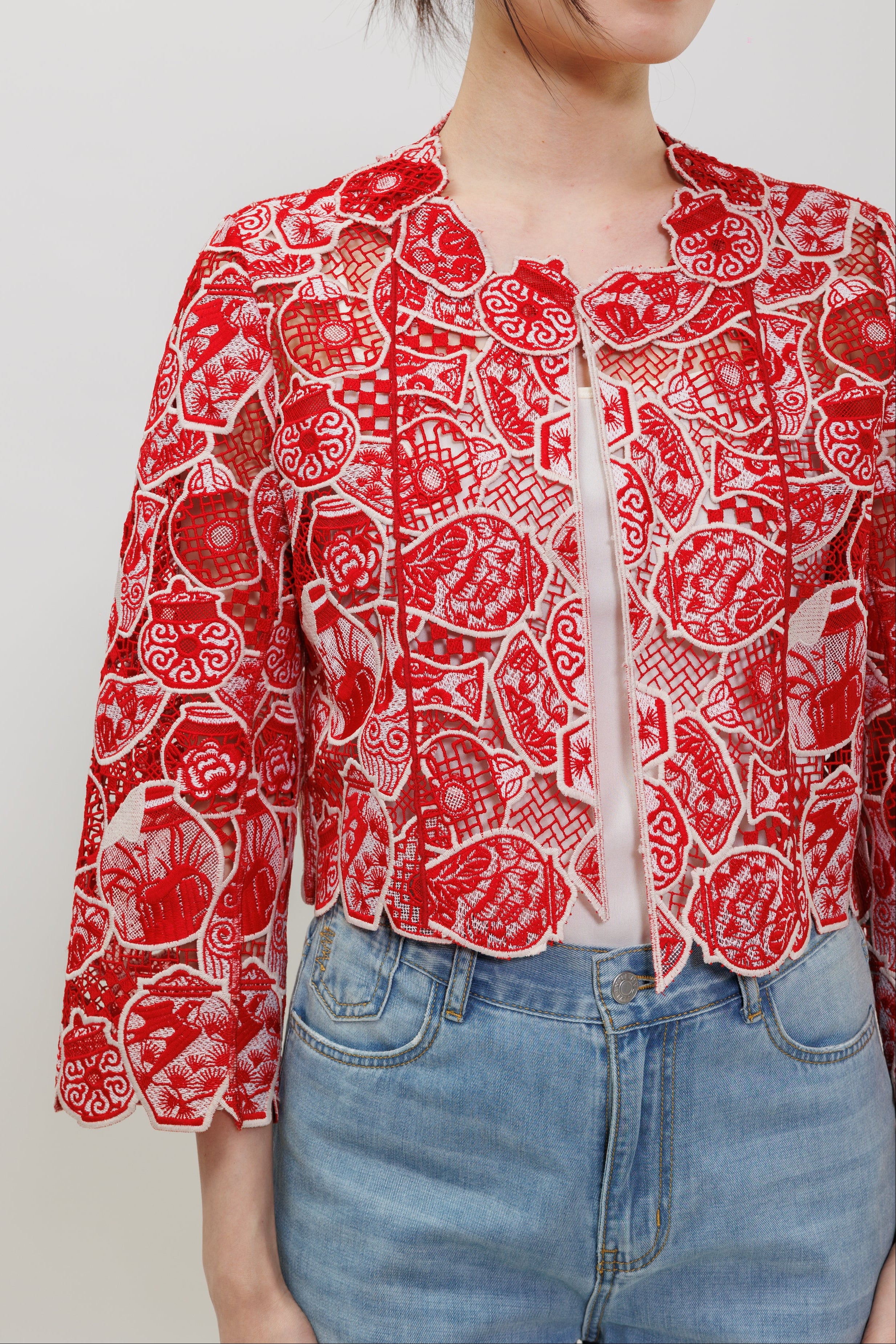 A woman wearing a red and white patterned cropped jacket with three-quarter sleeves, paired with light blue jeans, against a plain light gray background.
