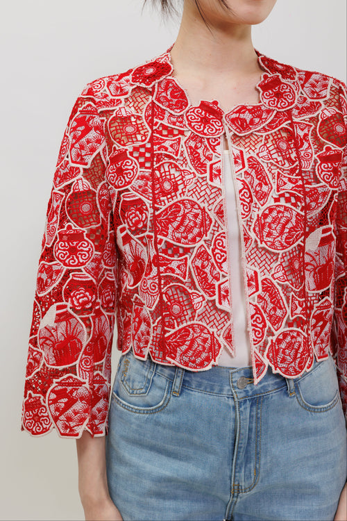 A woman wearing a red and white patterned cropped jacket with three-quarter sleeves, paired with light blue jeans, against a plain light gray background.