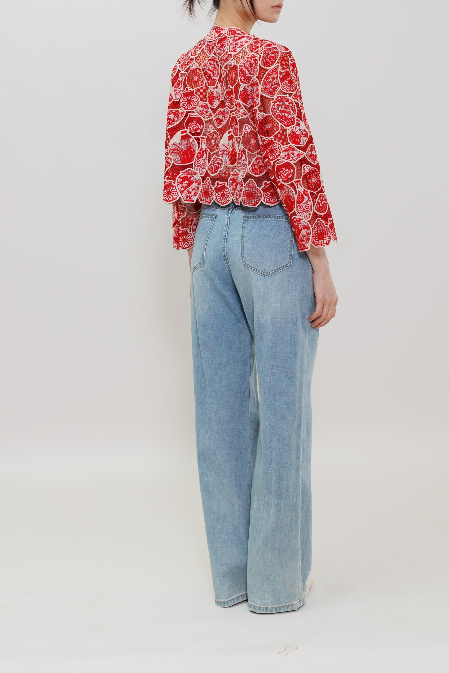 A woman stands in profile wearing a red and white floral long-sleeve blouse with light blue wide-leg jeans against a plain light gray studio background.