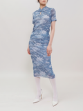 A model wearing a short-sleeved blue and white patterned cheongsam dress, paired with white tights and white pointed-toe heels, standing against a plain light gray studio background.