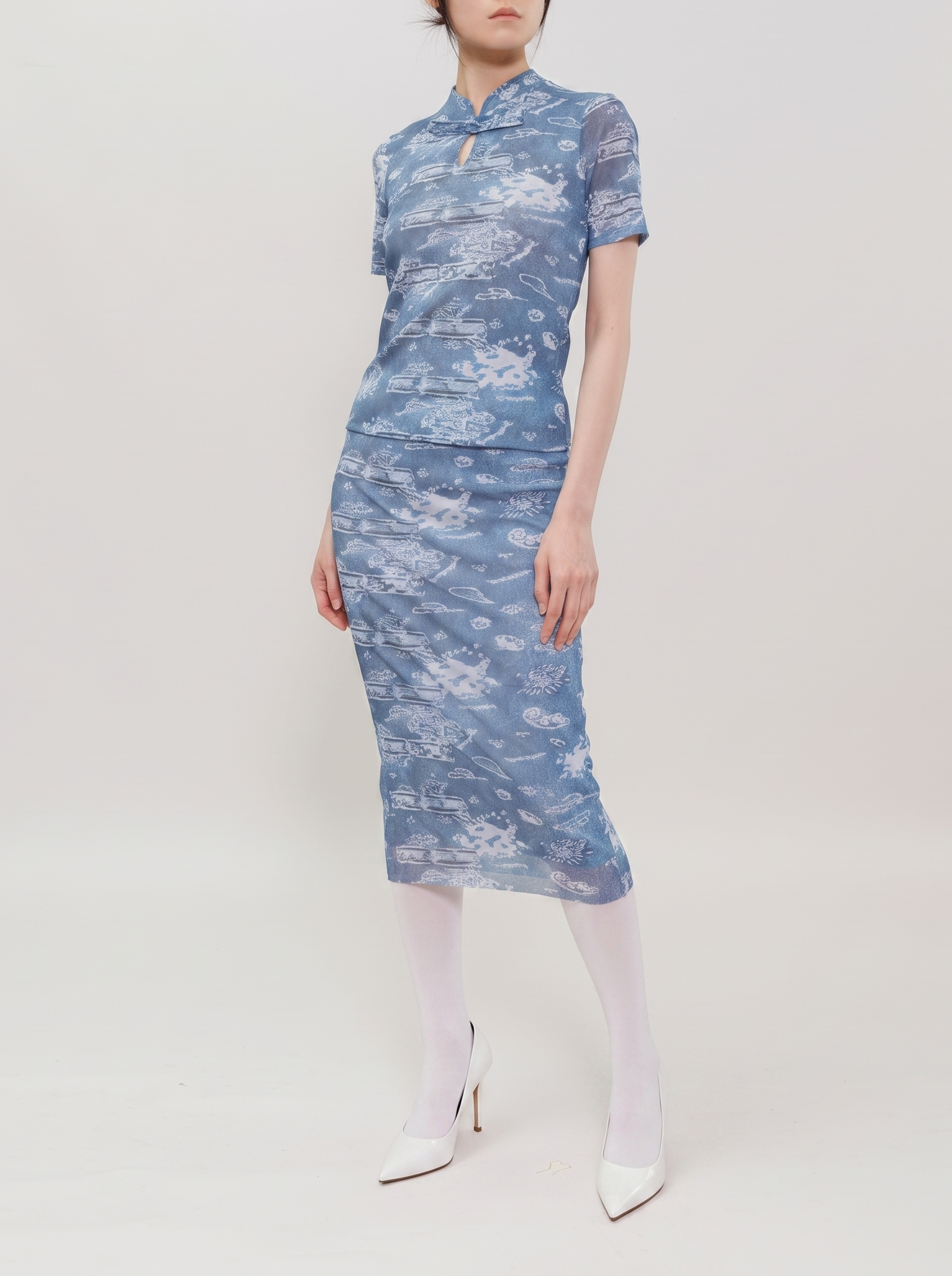 A model wearing a short-sleeved blue and white patterned cheongsam dress, paired with white tights and white pointed-toe heels, standing against a plain light gray studio background.