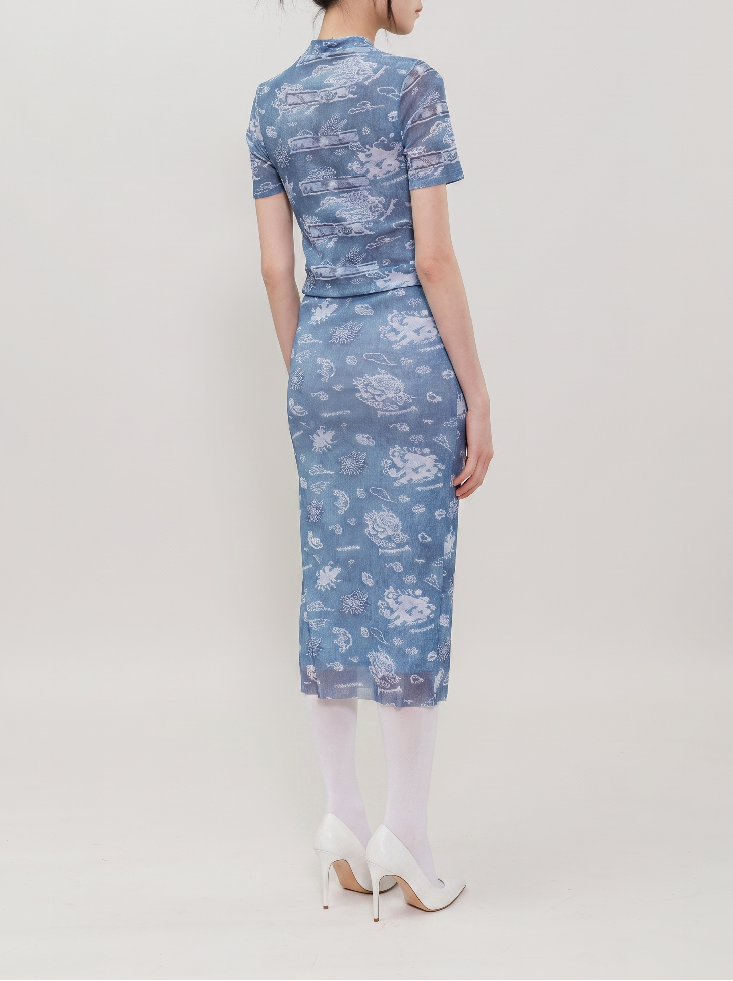 A woman is shown from the back wearing a short-sleeve blue dress with a white floral and cloud pattern, paired with white tights and white high heels, against a plain light gray studio background.