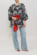 A model stands against a light gray studio background wearing a black and gray abstract patterned long-sleeve top, light blue wide-leg jeans, and red pointed shoes, while holding a red clutch with tassels.
