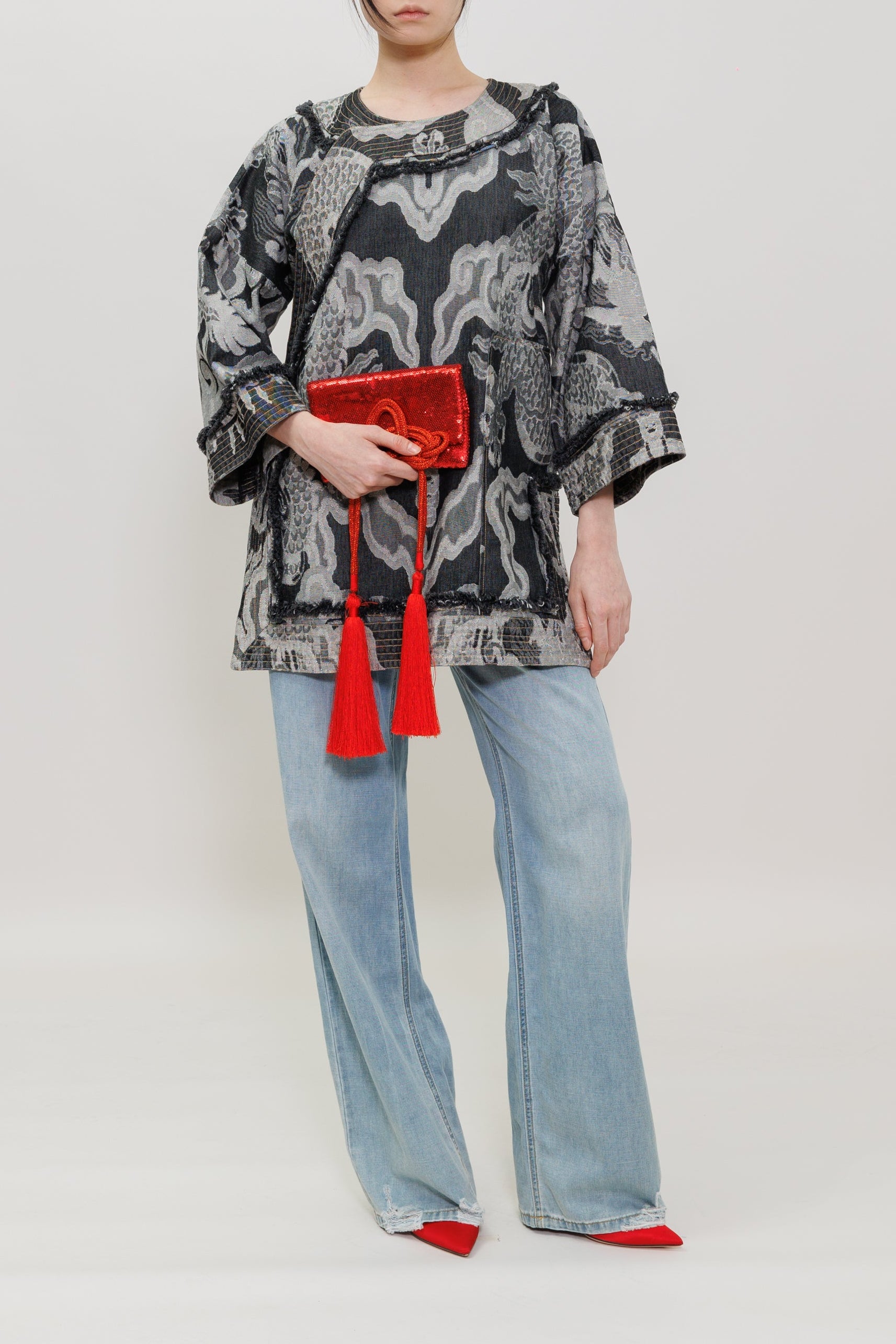 A model stands against a light gray studio background wearing a black and gray abstract patterned long-sleeve top, light blue wide-leg jeans, and red pointed shoes, while holding a red clutch with tassels.