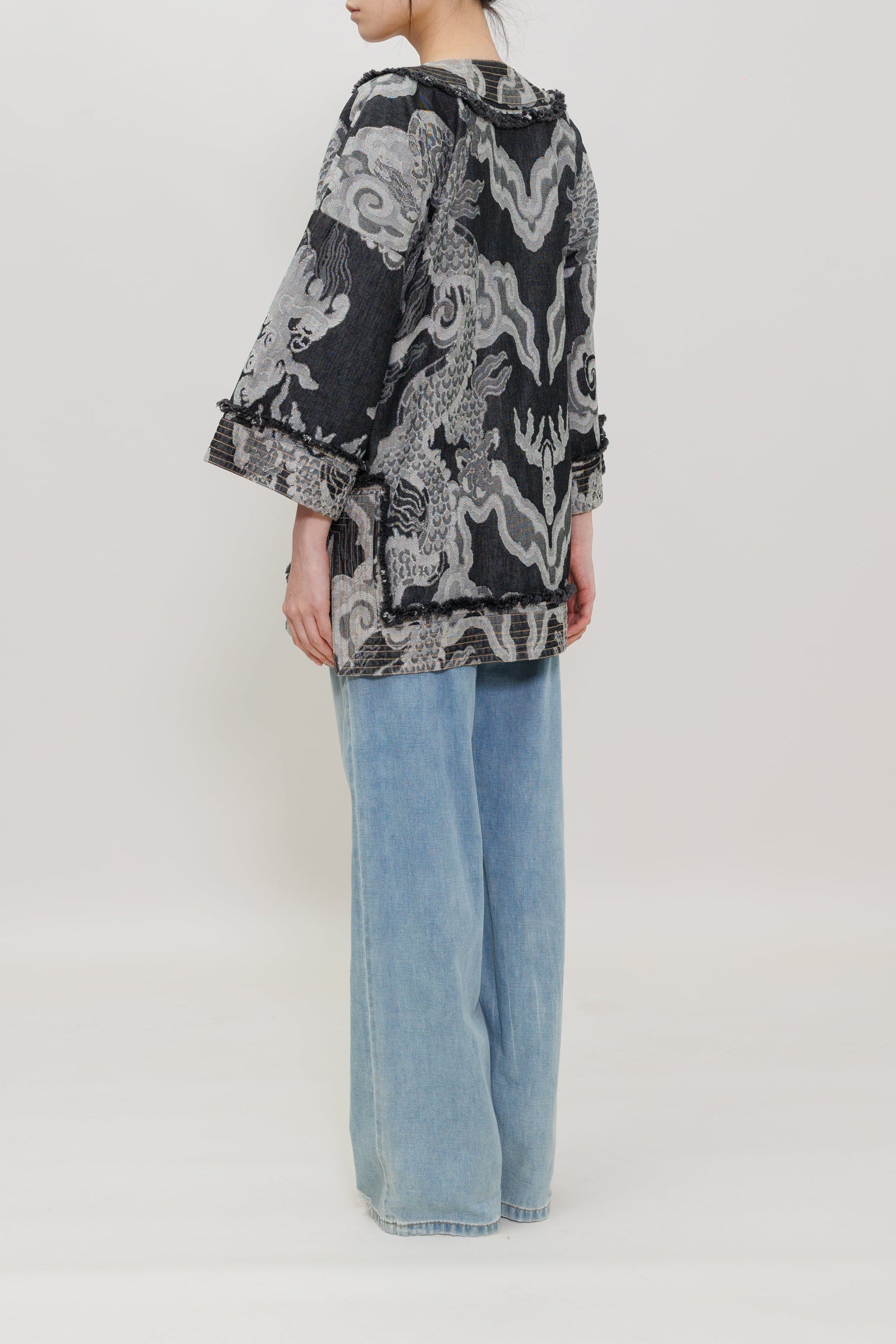 A model stands in profile wearing a black and white abstract patterned cardigan with wide three-quarter sleeves, paired with light blue wide-leg jeans, against a plain white studio background.