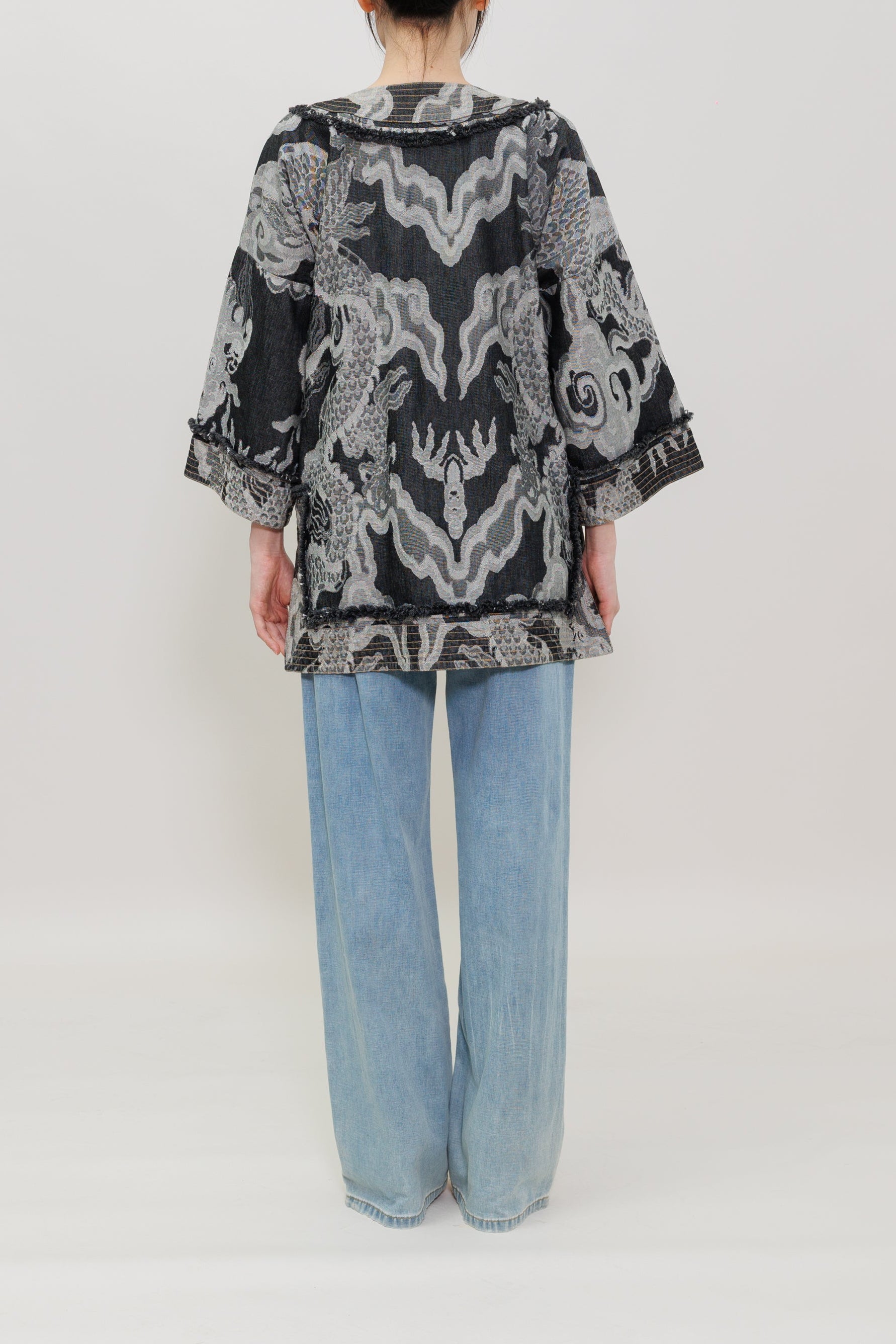 A person viewed from the back wearing a loose black and white patterned top with wide light blue jeans, standing against a plain light gray studio background.