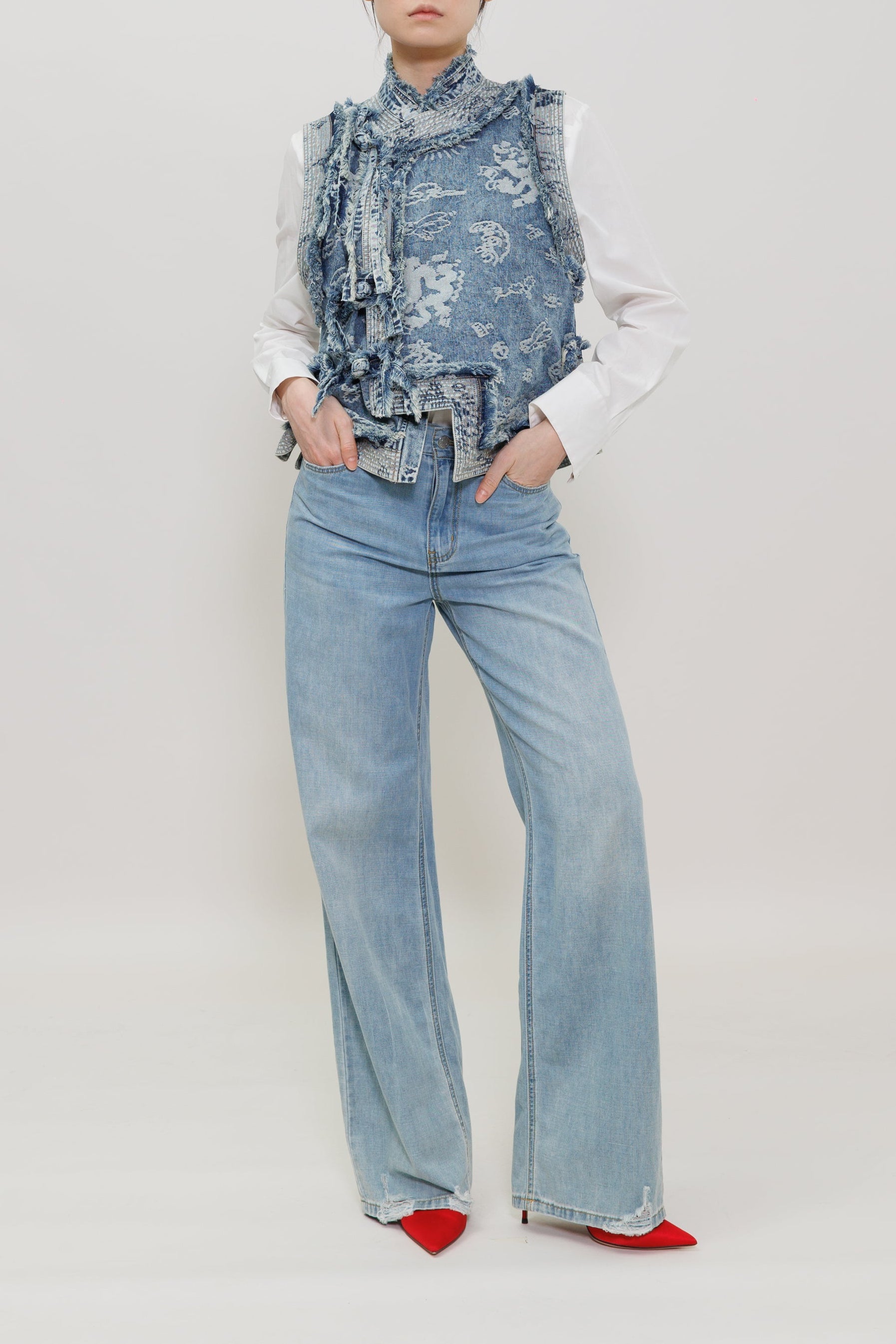 A model stands against a plain light gray studio background wearing a light blue denim vest over a white ruffled high-neck blouse, light-wash wide-leg jeans, and bright red pointed shoes.