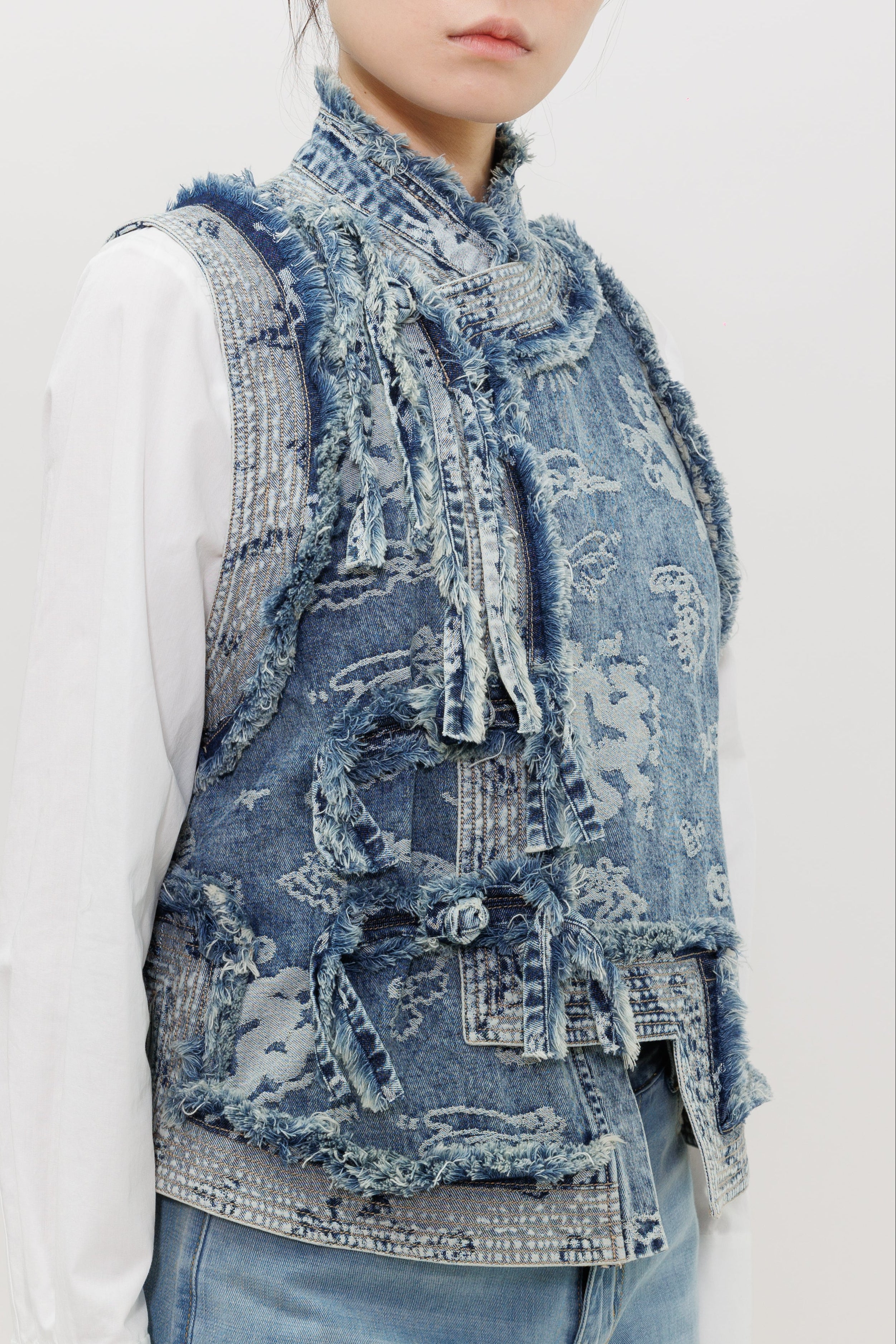 A close-up of a model wearing a distressed, layered blue denim vest over a white long-sleeve shirt and light blue jeans, against a plain light gray background.