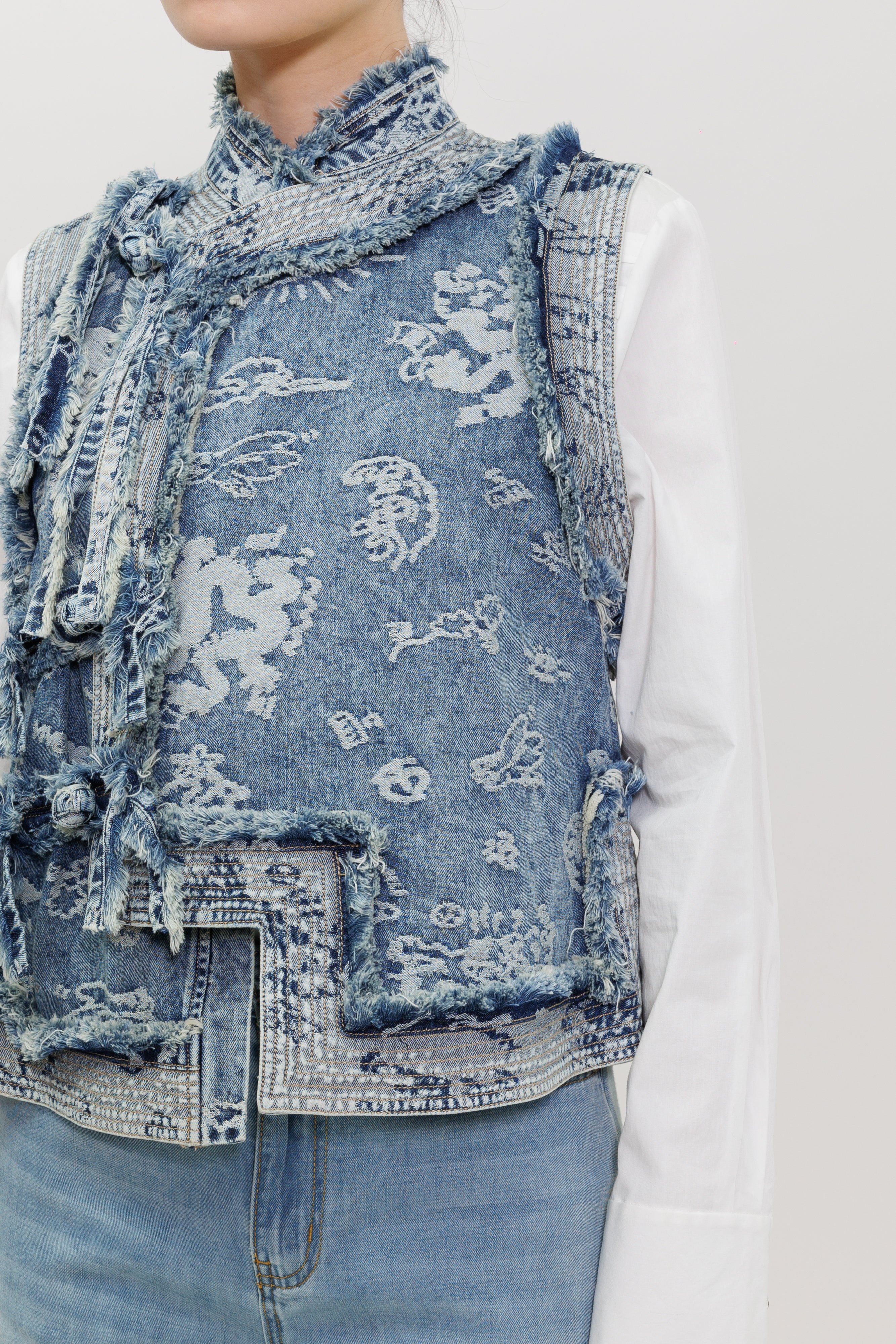 A close-up of a model wearing a distressed blue denim vest with white abstract patterns, layered over a white long-sleeve shirt and light-wash jeans.