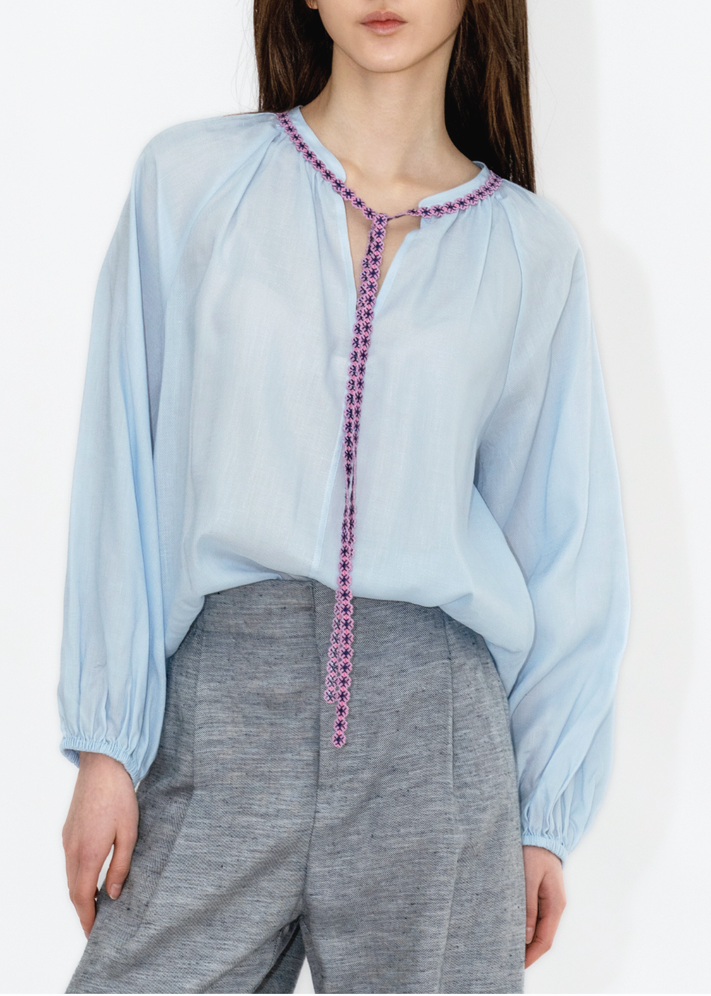 A woman wearing a light blue, fine twill blouse with long sleeves and a round neckline with a decorative purple and pink beaded trim, paired with gray trousers.