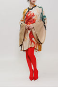 A woman stands against a plain light gray studio background wearing a loose, long-sleeved beige top with a bold red, orange, and green dragon print, paired with bright red tights and red high heels.