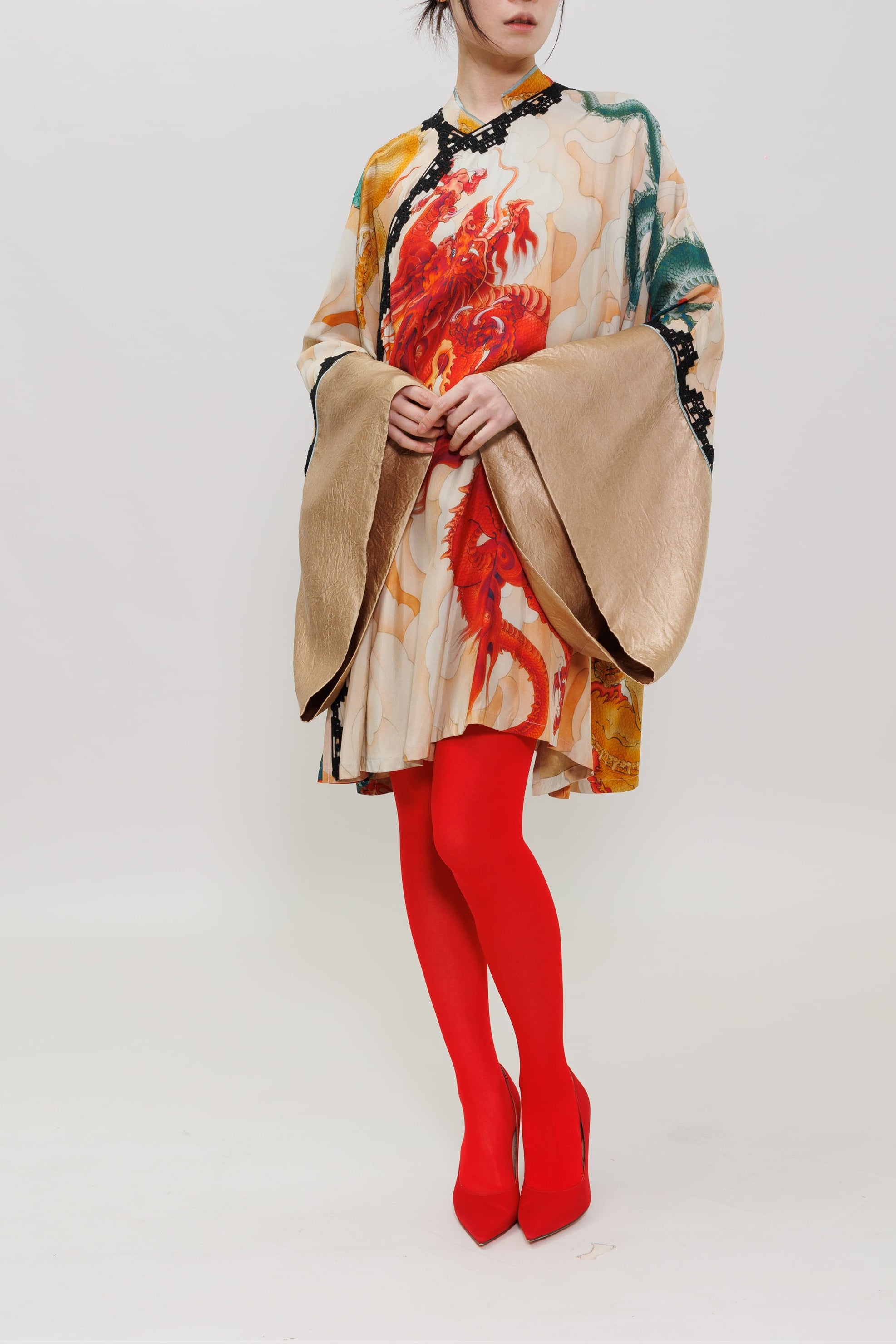 A woman stands against a plain light gray studio background wearing a loose, long-sleeved beige top with a bold red, orange, and green dragon print, paired with bright red tights and red high heels.