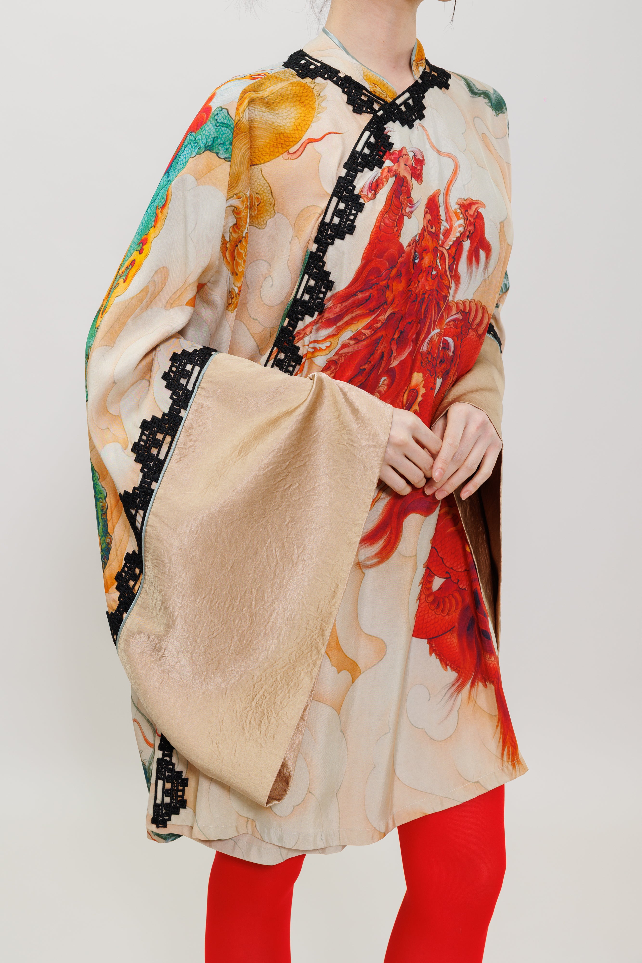 A model wearing a beige silk-like tunic with a vibrant red, orange, and teal abstract print, featuring black lace trim and wide draped sleeves, paired with bright red leggings against a light gray background.