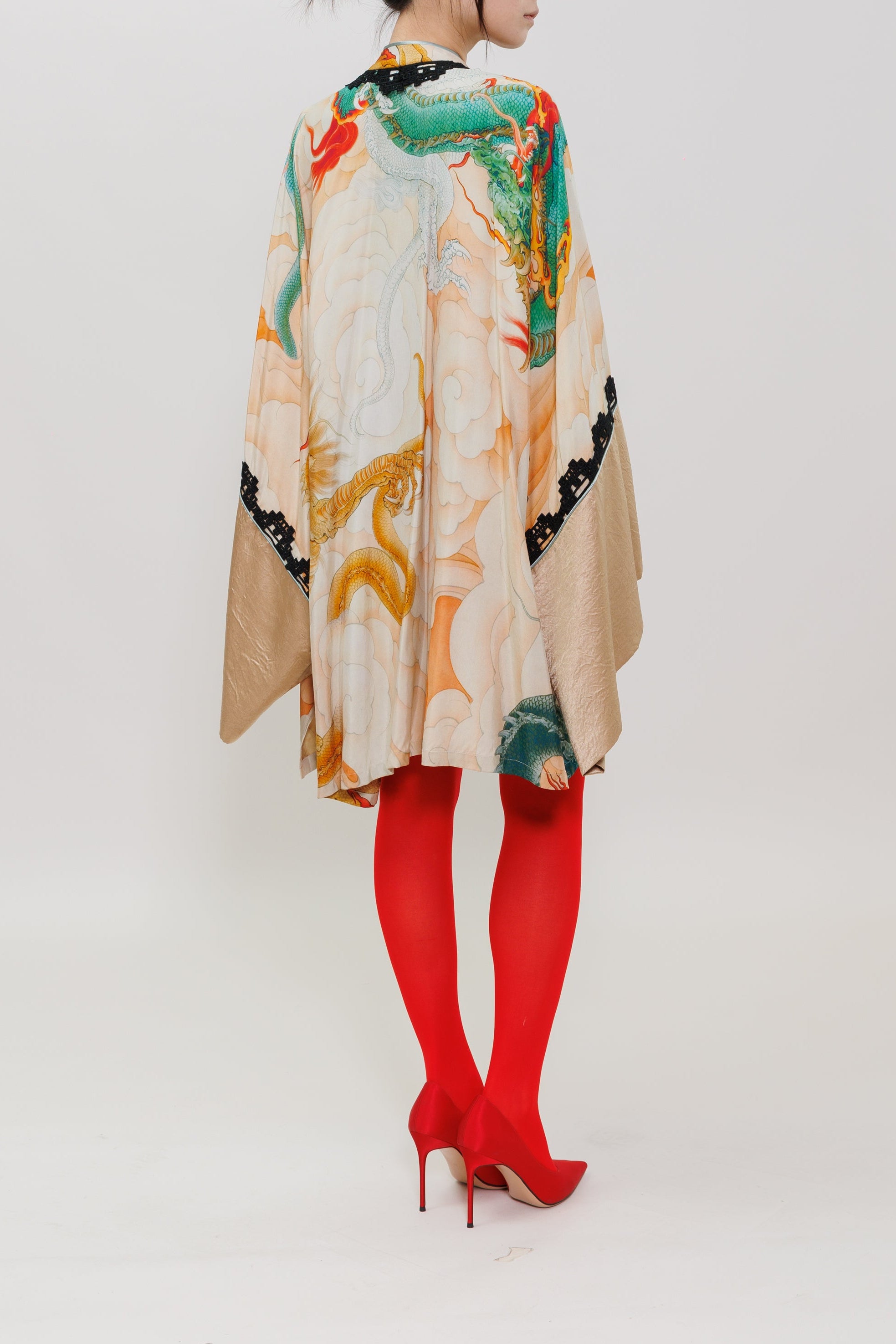 A woman stands in profile wearing a beige kimono-style poncho with a colorful green, red, and gold dragon print, paired with bright red tights and red high heels, against a plain white studio background.