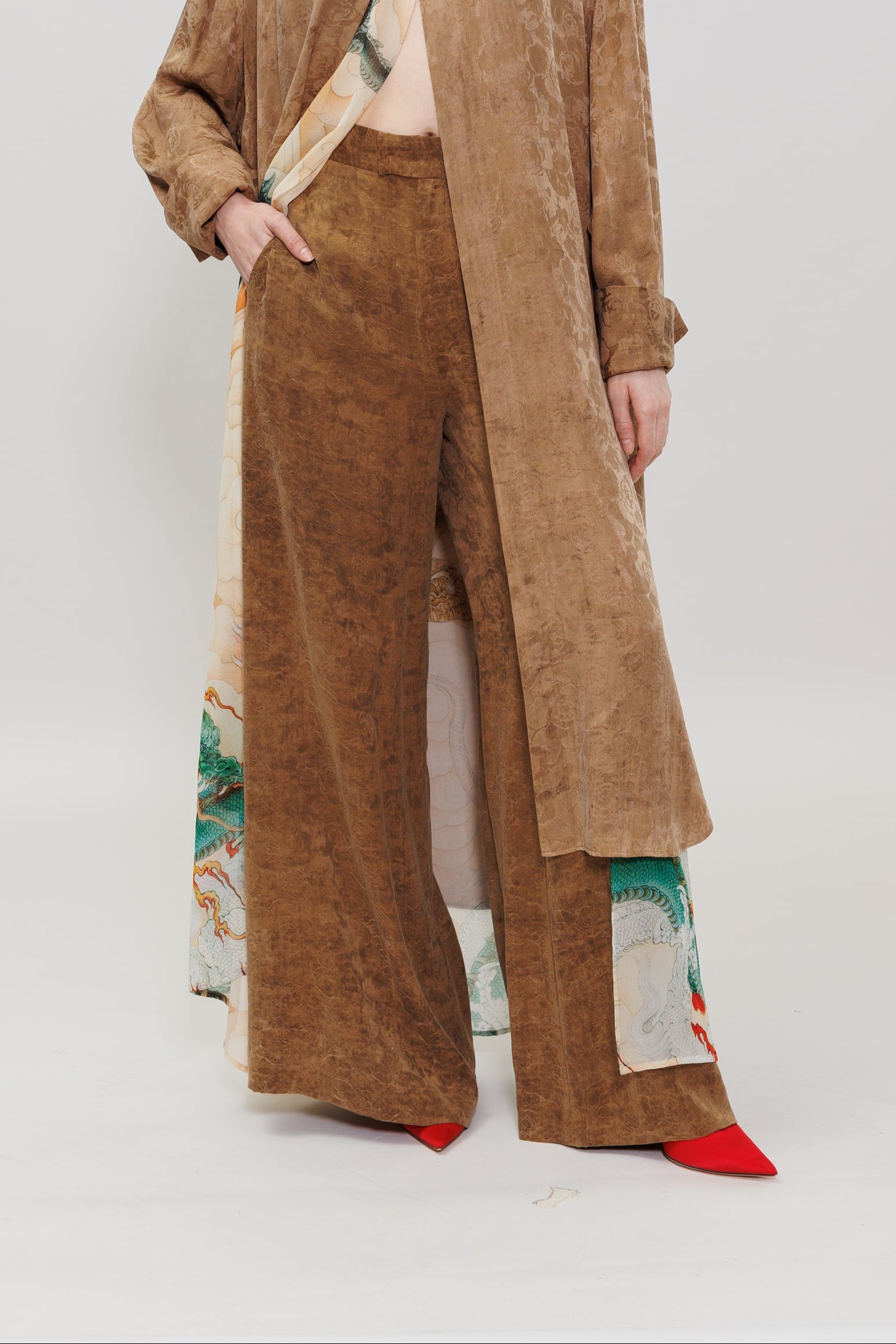 A model stands against a plain light gray studio background wearing a layered fashion look: a long tan suede-like overshirt over a light top, wide-leg brown pants, and bright red pointed shoes. The pants and lower hem of the outer layer show a visible multicolor print along the side.