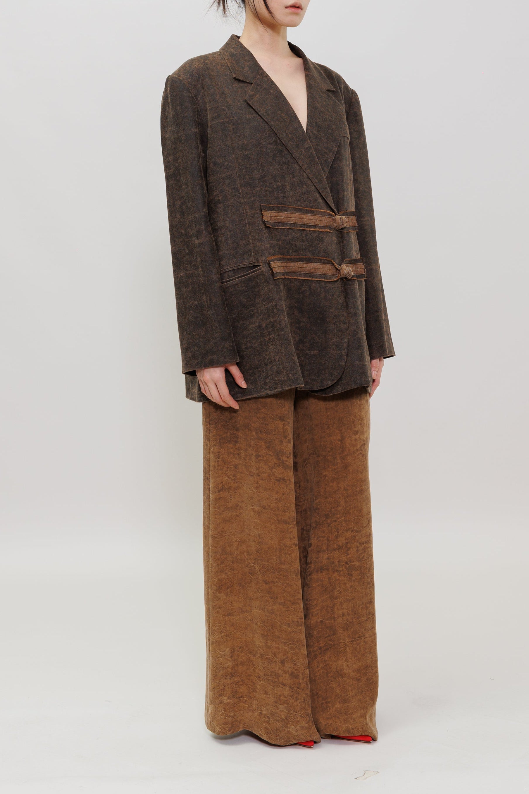 A model stands against a plain light gray studio background wearing a dark brown textured blazer with matching tie details and wide-leg brown pants.