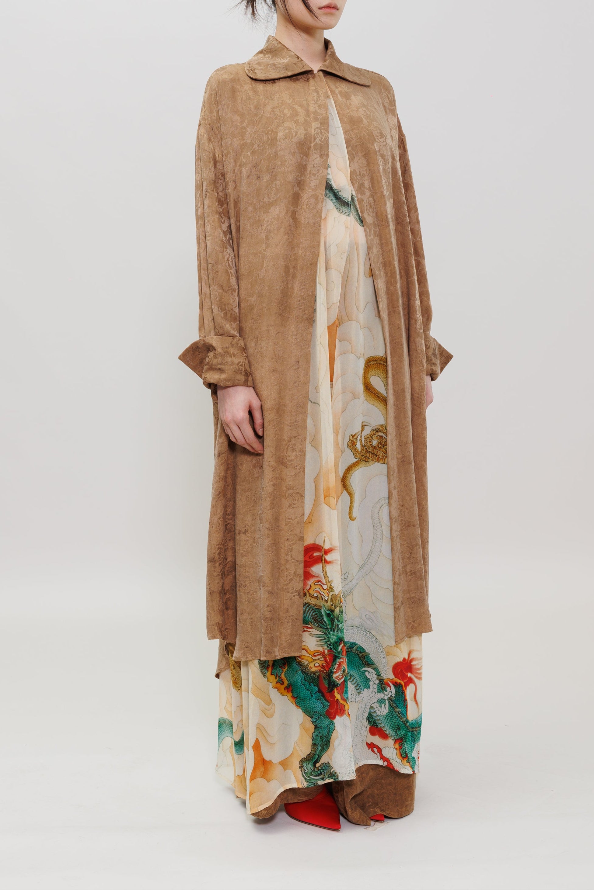 A model stands against a plain light gray studio background wearing a long tan suede-like coat over a cream dress with a colorful dragon print and red shoes.