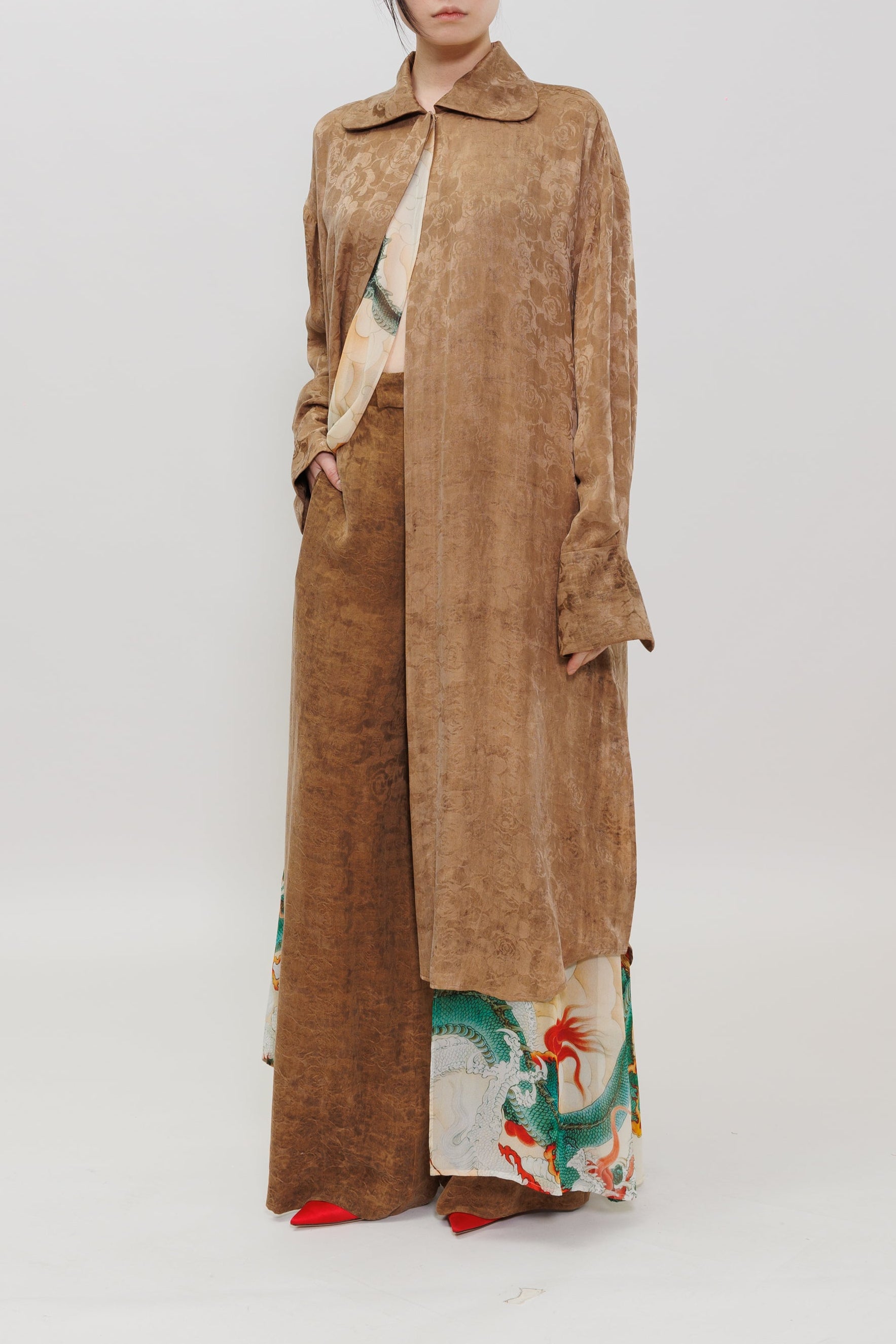 A model stands against a plain light gray studio background wearing a long, loose brown coat over a cream and green patterned kimono-style dress with wide legs.