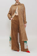A model stands against a plain light gray studio background wearing a layered fashion look: a long tan suede-like overshirt over a light top, wide-leg brown pants, and bright red pointed shoes. The pants and lower hem of the outer layer show a visible multicolor print along the side.