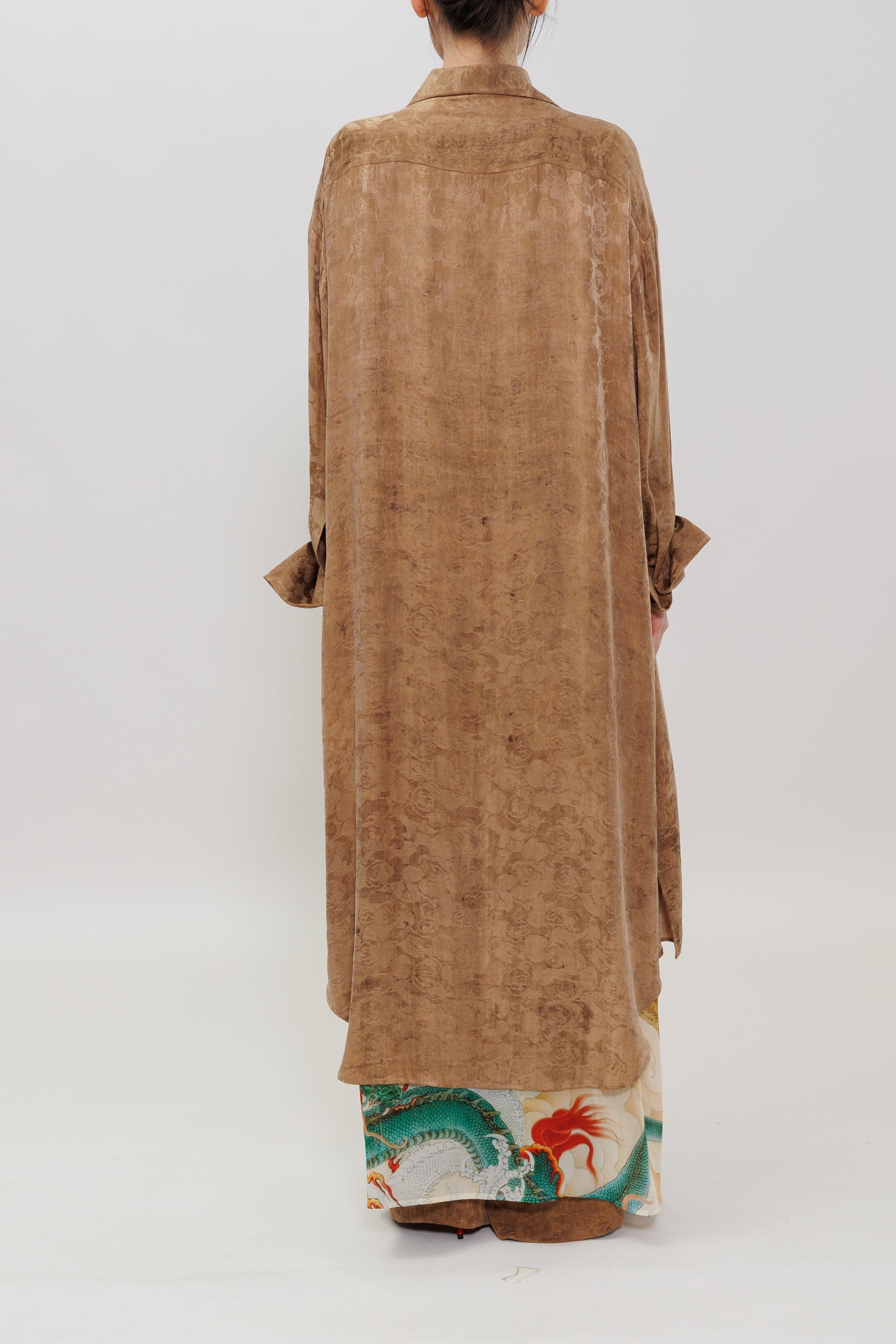 A model viewed from behind wearing a long, loose-fitting brown jacket over a colorful patterned skirt, set against a plain light gray studio background.