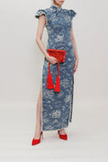 A woman modeling a blue and white floral print cheongsam dress, holding a red clutch with red tassels, and wearing red pointed-toe heels against a plain light gray studio background.