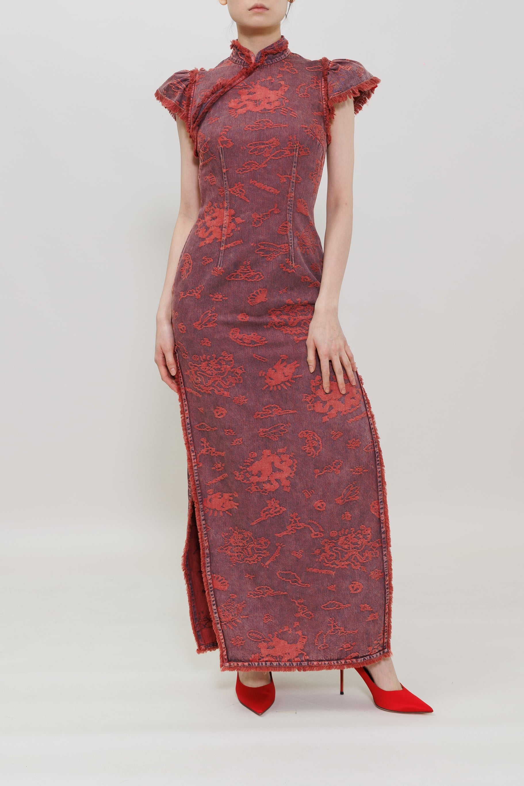 A woman modeling a long red and black floral-patterned cheongsam dress with red high heels, standing against a plain light gray studio background.