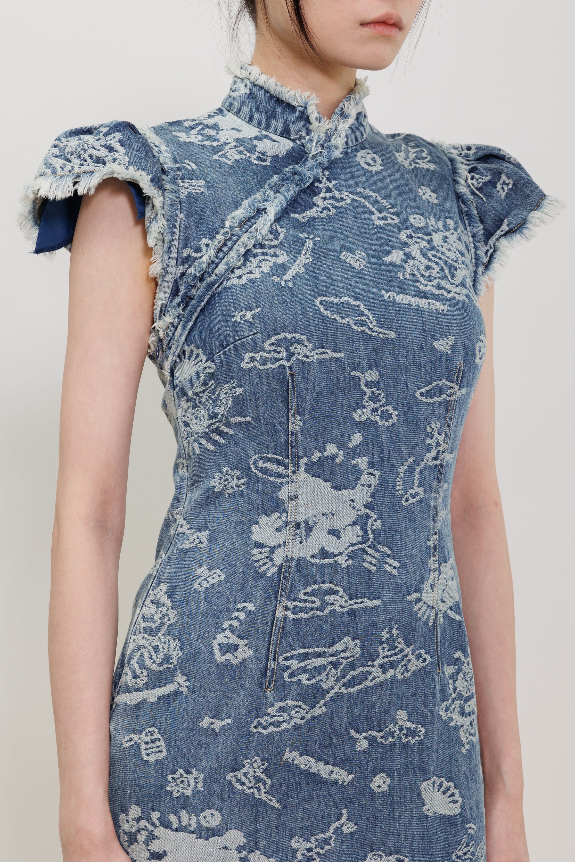 A model wearing a blue denim cheongsam dress with white printed cloud and animal motifs, featuring ruffled cap sleeves and a high collar, set against a plain light gray background.