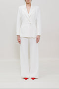 A model stands against a seamless white studio background wearing a white tailored blazer with a matching tie belt and wide-leg trousers, paired with bright red pointed shoes.