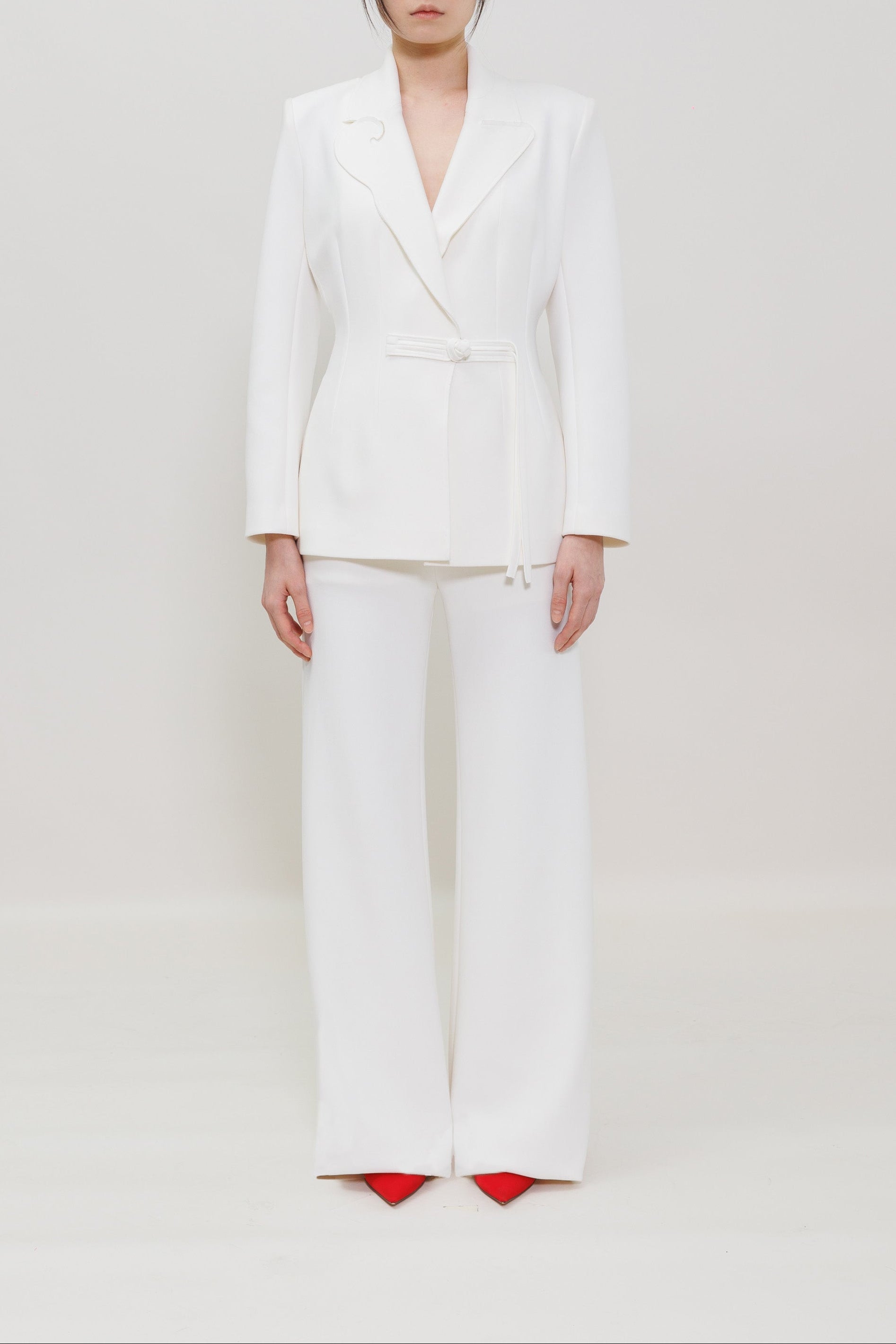 A model stands against a seamless white studio background wearing a white tailored blazer with a matching tie belt and wide-leg trousers, paired with bright red pointed shoes.