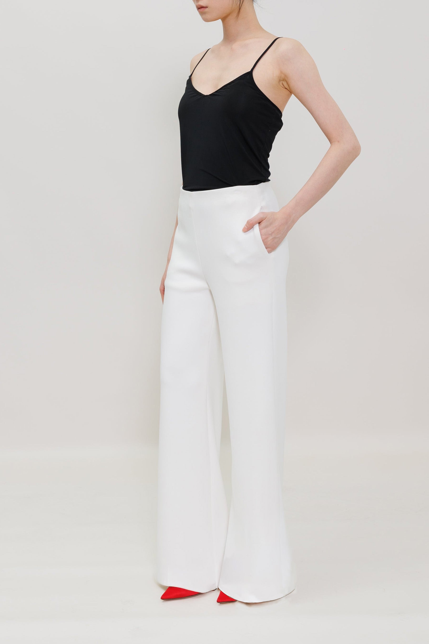 A model wearing a black camisole top and high-waisted white wide-leg trousers, standing against a light gray studio background.