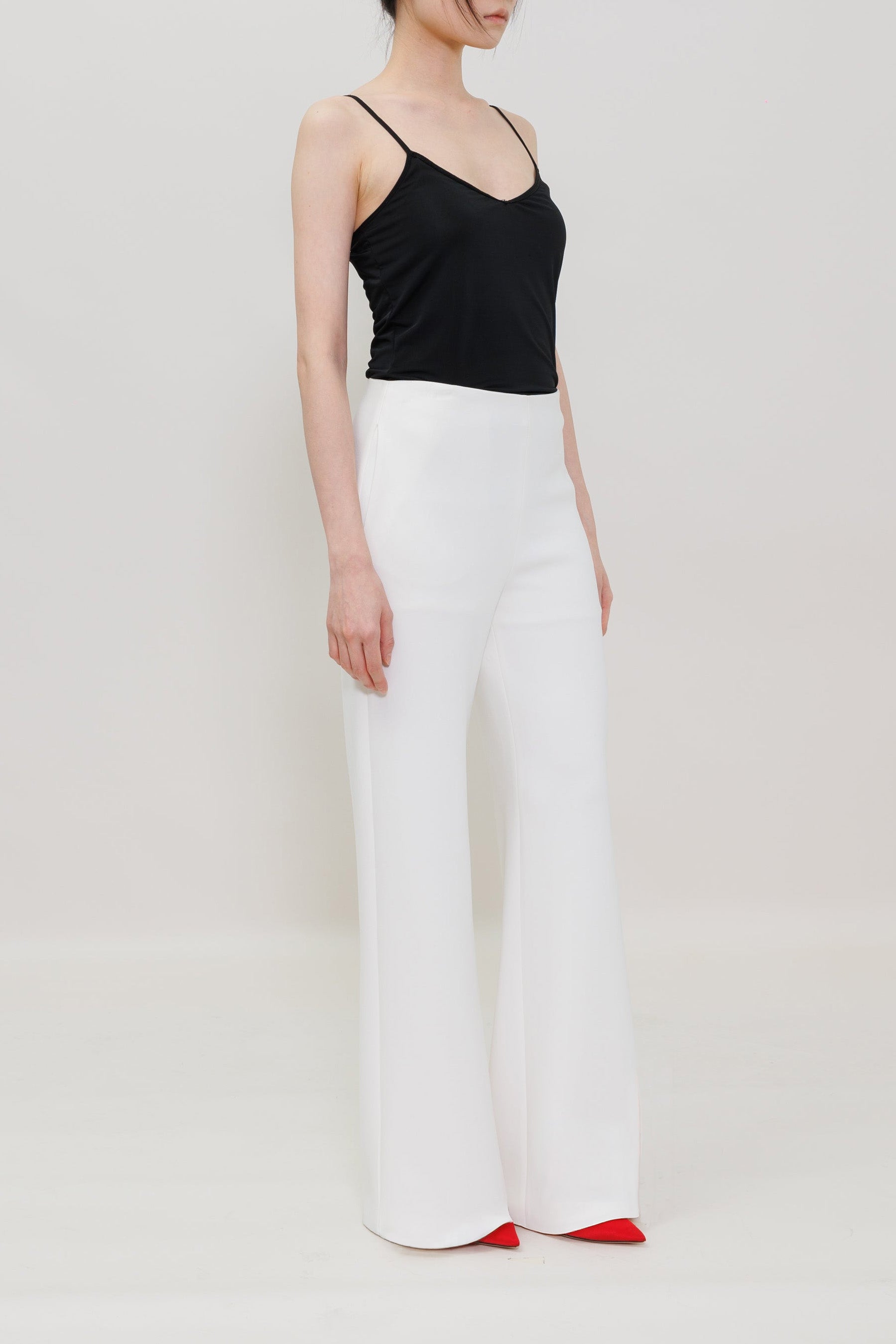 A model wearing a black spaghetti-strap camisole top and high-waisted white wide-leg trousers, paired with red pointed-toe heels, against a plain light gray studio background.