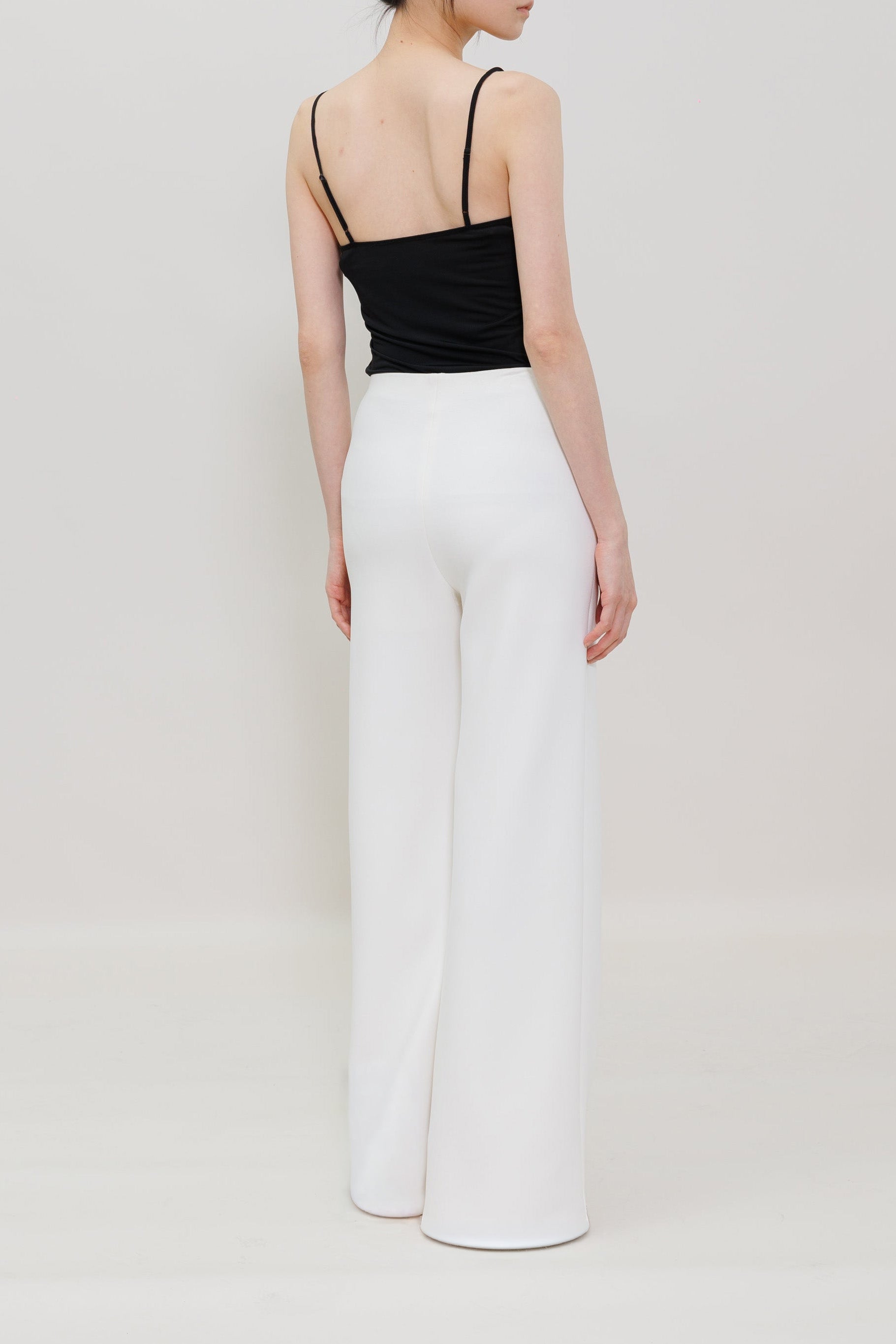 A model shown from the back wearing a black spaghetti-strap camisole top and high-waisted white wide-leg trousers, set against a plain light gray studio background.