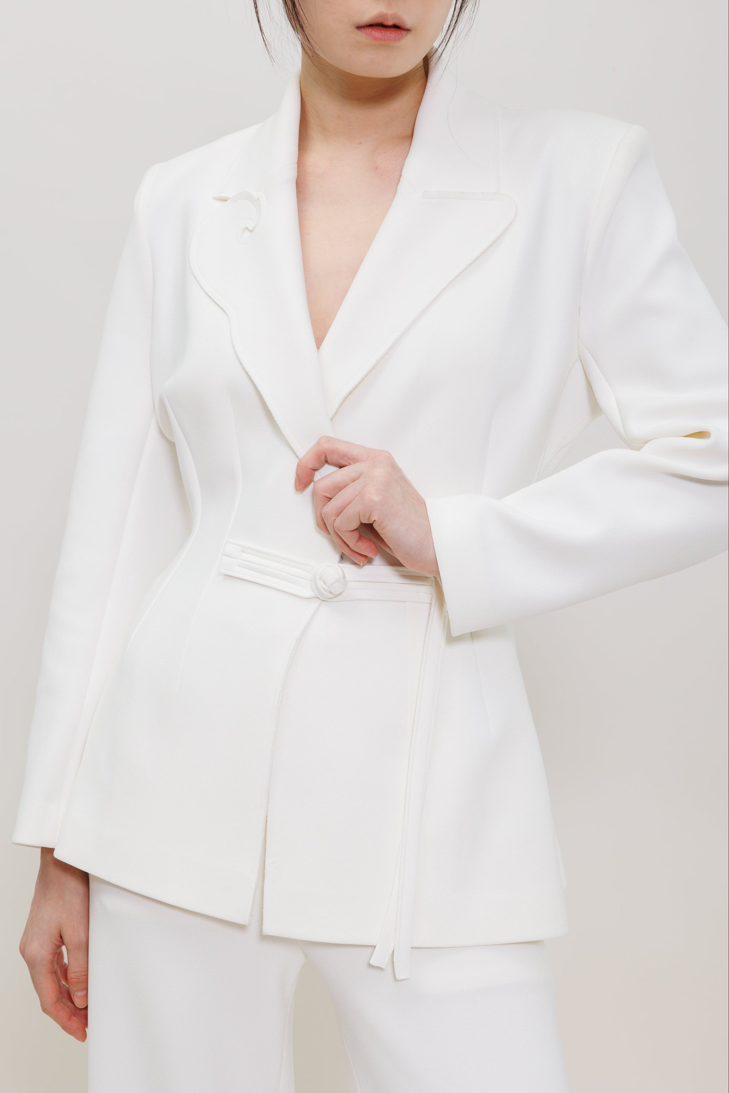 A model wearing a structured white blazer with a wrap-style tie belt at the waist, paired with matching white trousers, against a plain light background.