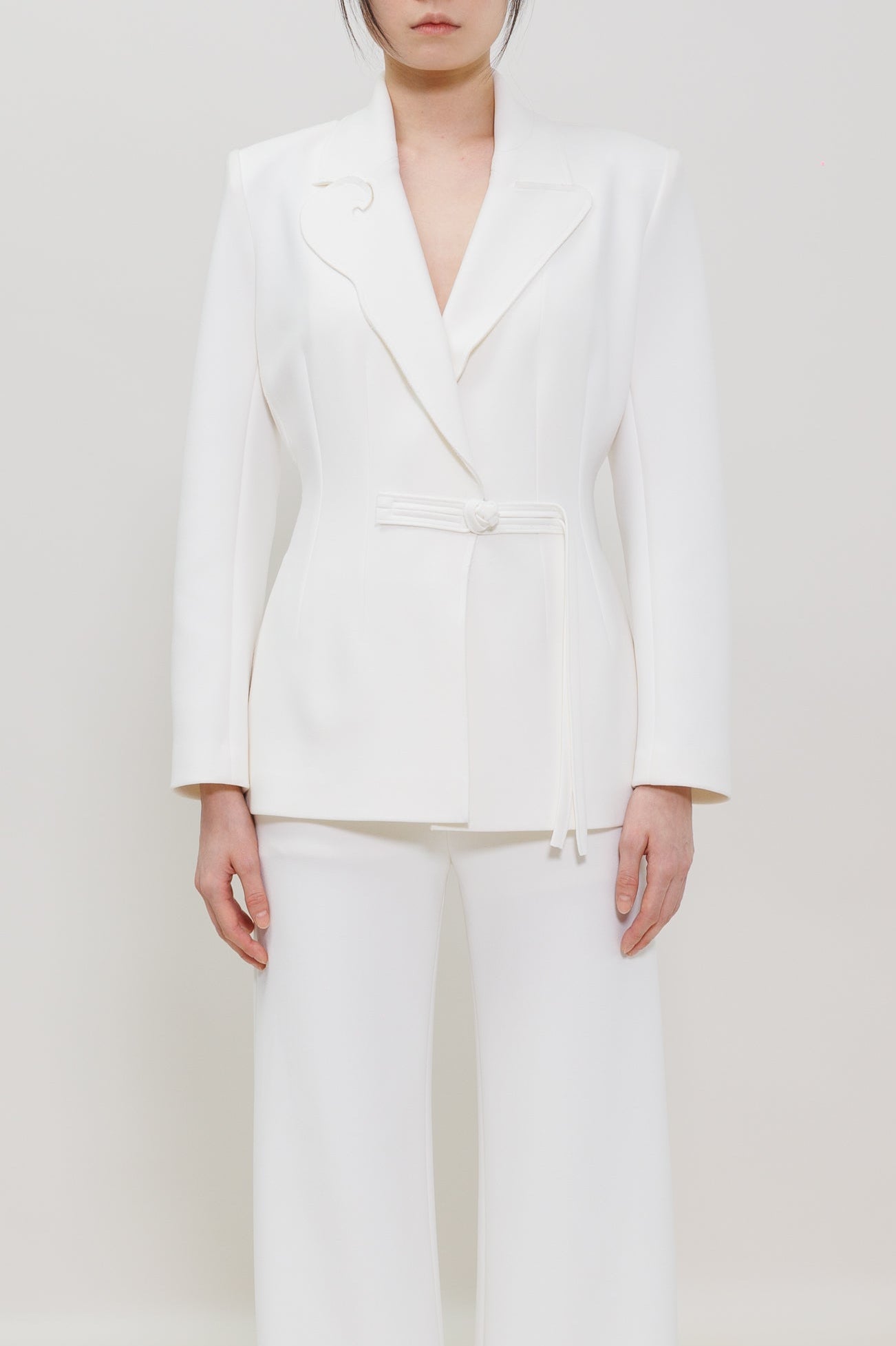A model stands against a seamless white studio background wearing a white tailored blazer with a matching tie belt and wide-leg trousers, paired with bright red pointed shoes.