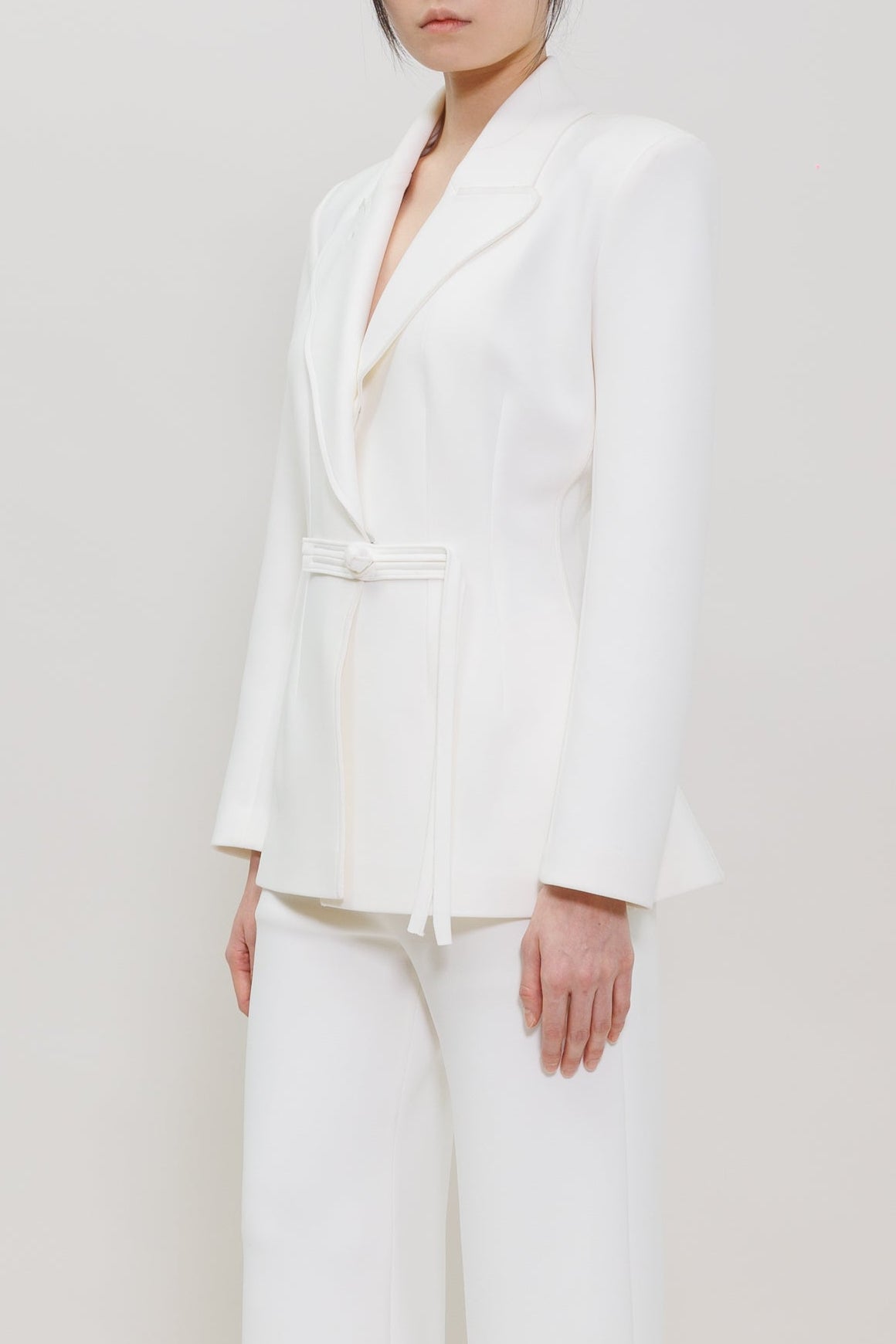 A model stands in a studio wearing a white tailored suit consisting of a belted wrap-style blazer and matching wide-leg trousers, paired with bright red pointed shoes against a plain light background.