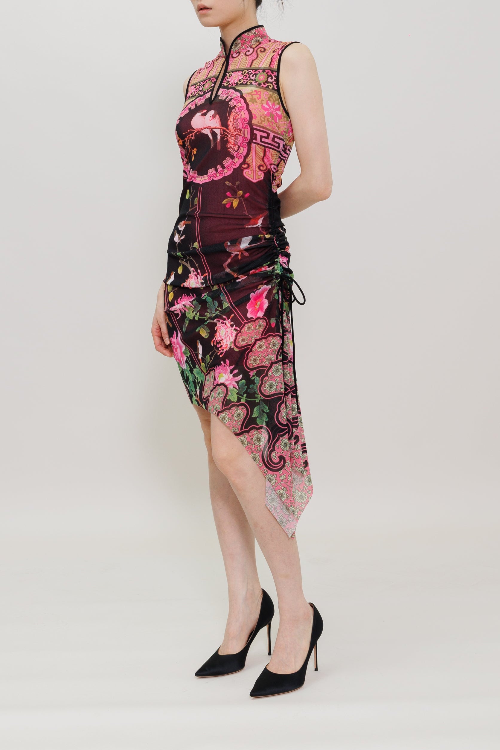 A model wearing a sleeveless pink and black floral embroidered cheongsam dress with black high heels, standing against a plain white studio background.