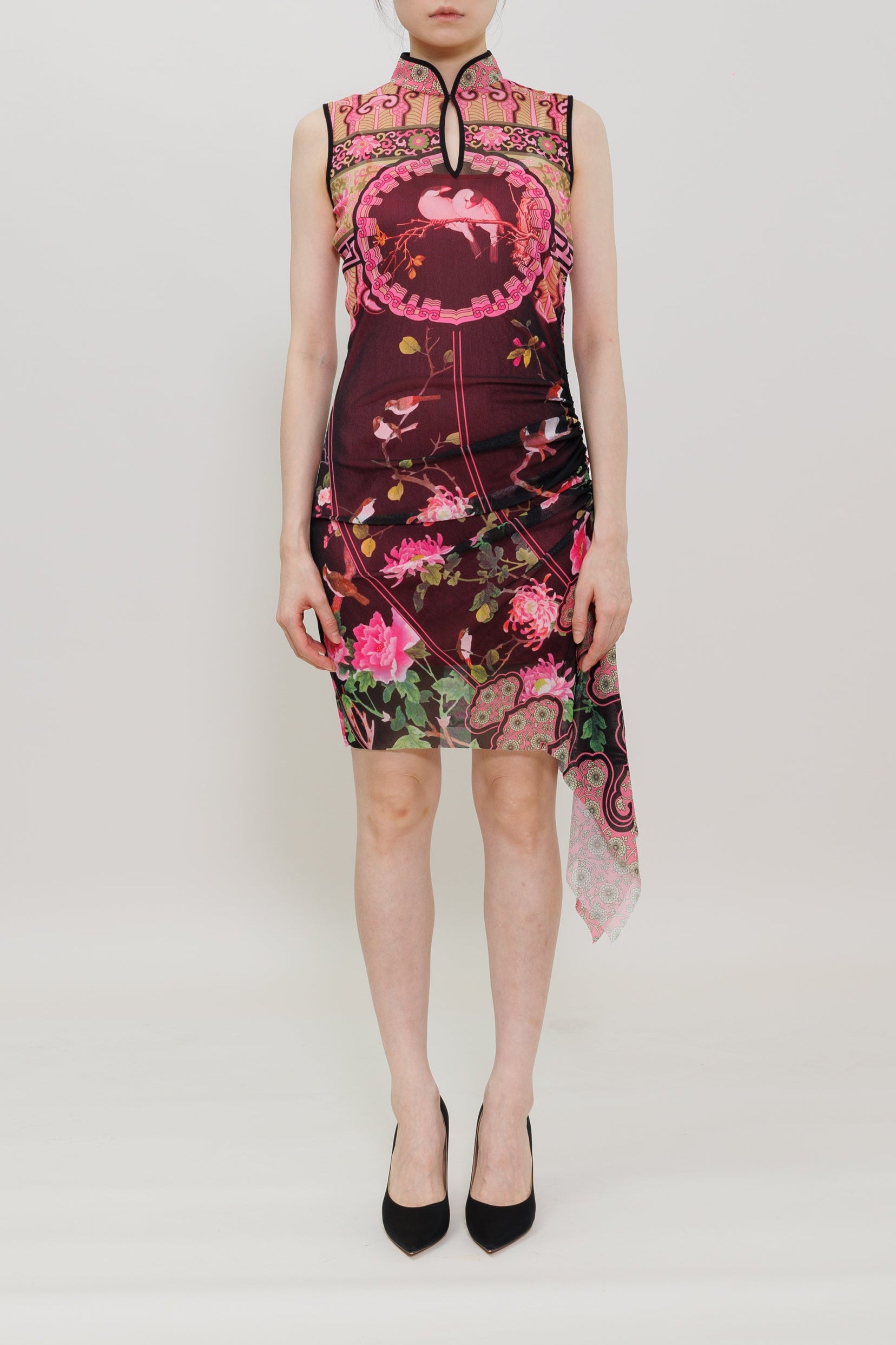 A model stands against a plain white studio background wearing a sleeveless dark pink and black floral dress with a high collar and black pointed heels.