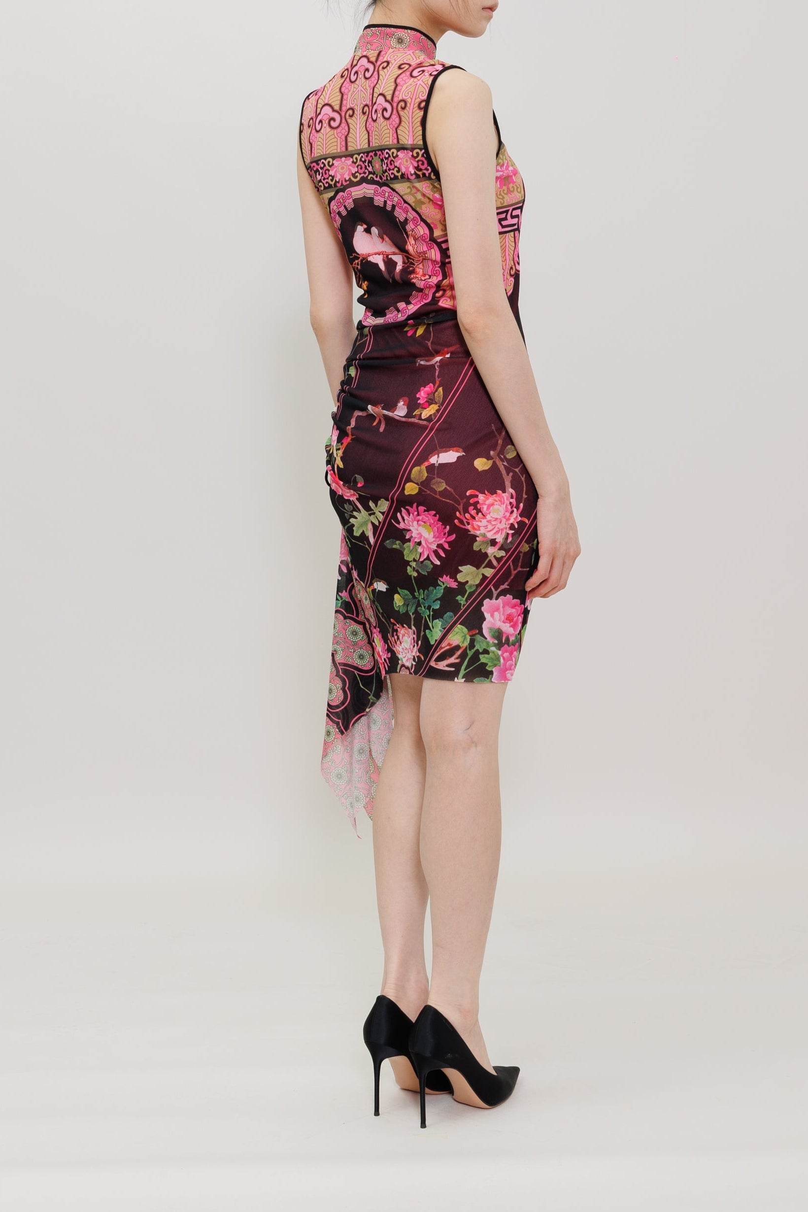 A woman stands in profile wearing a sleeveless, high-neck mini dress with a pink and green floral print on a dark background, paired with black high heels, against a plain light gray studio backdrop.
