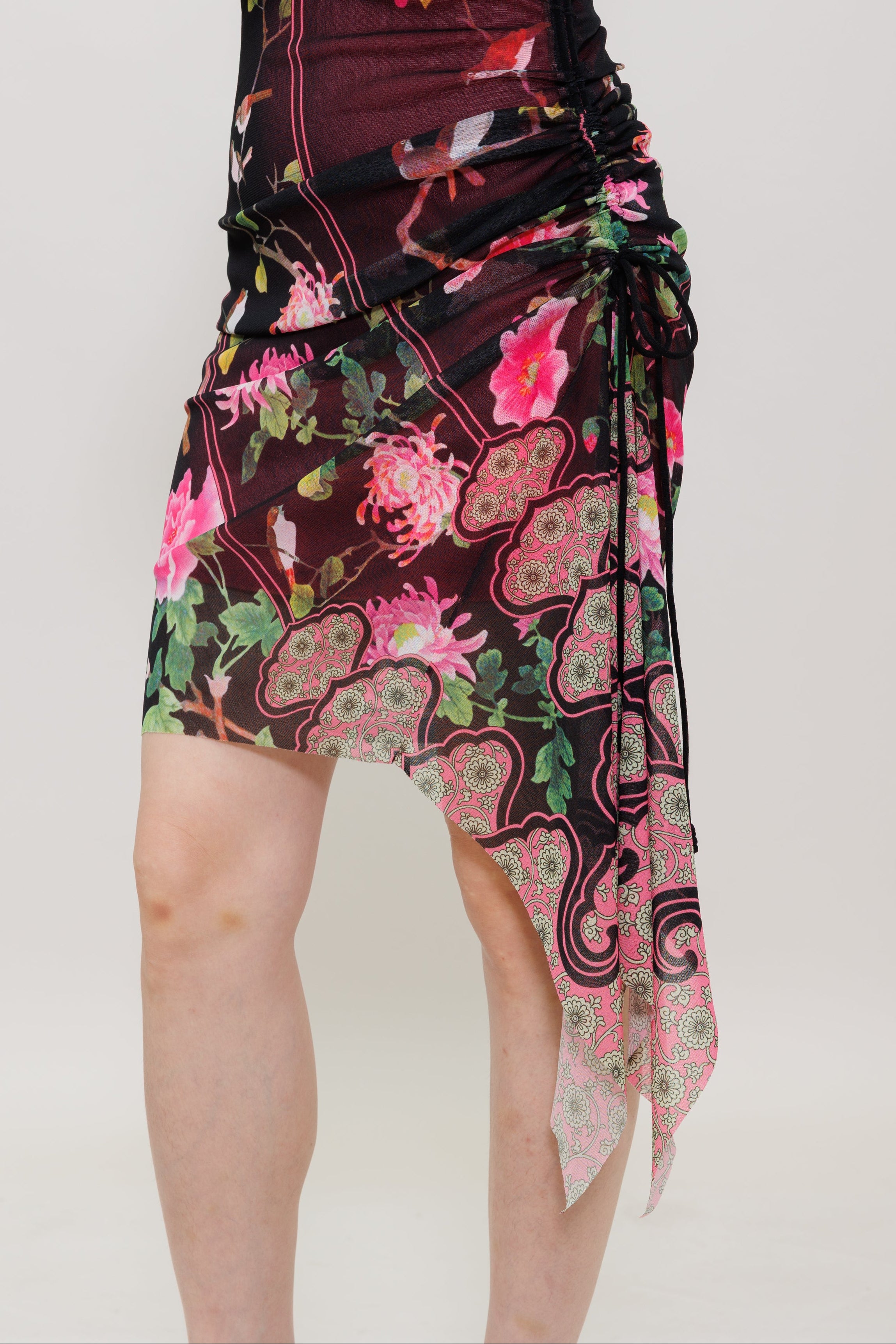 A model wearing a ruched wrap-style dress with a dark burgundy base and a vibrant pink, green, and black floral paisley print, shown against a plain light gray studio background.