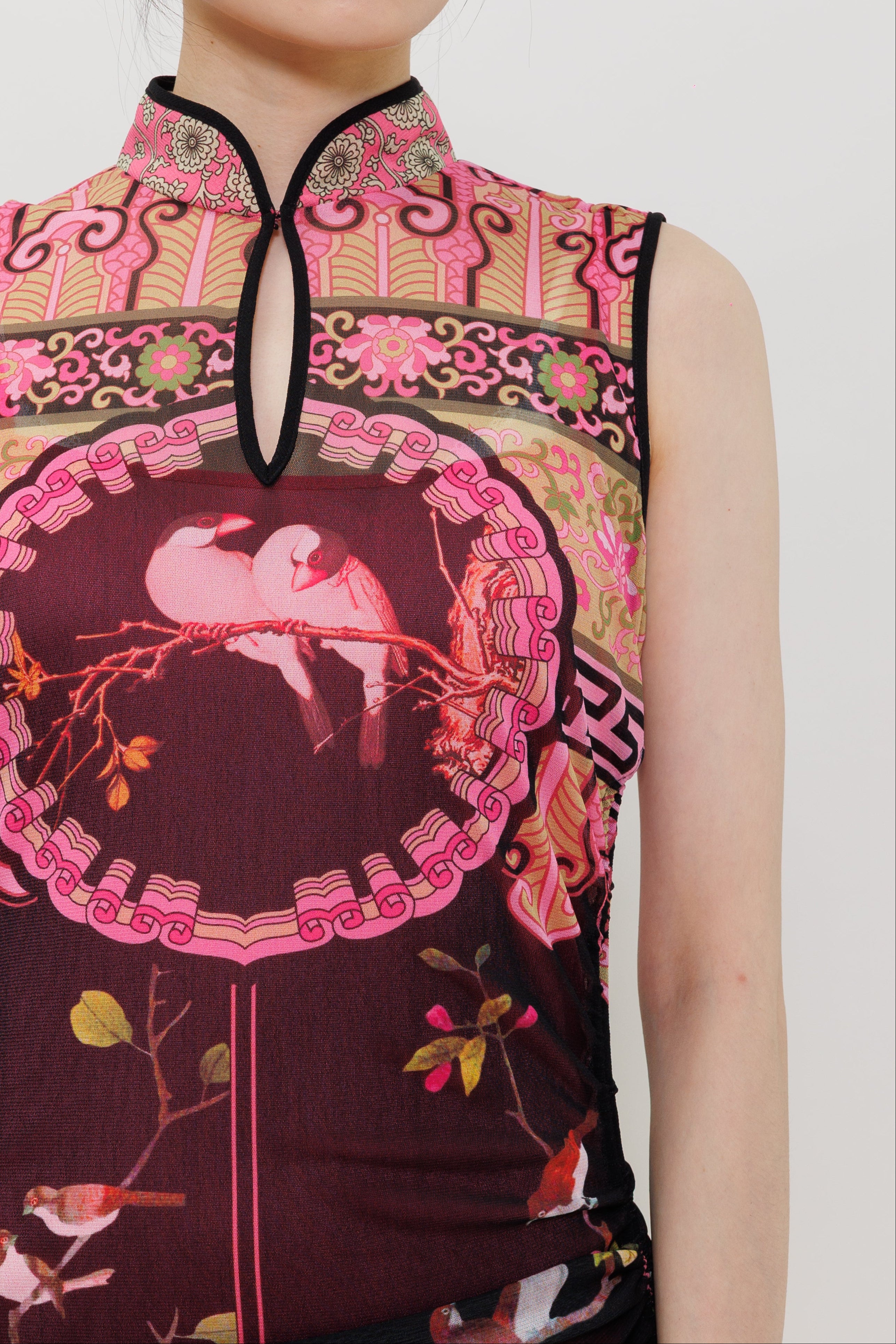 A close-up of a model wearing a sleeveless cheongsam-style top with a pink and dark plum print featuring birds and ornate traditional patterns, set against a plain light gray background.